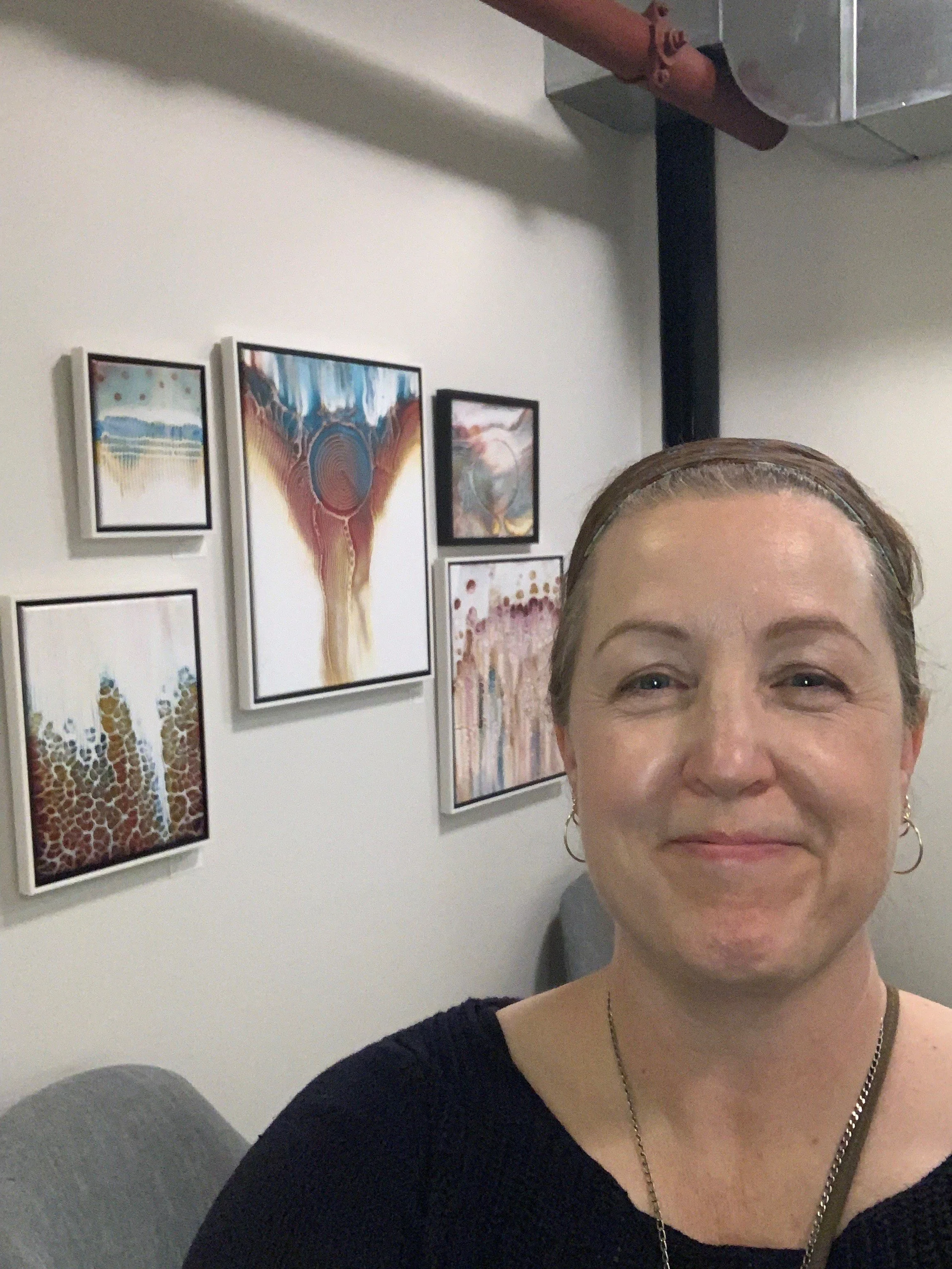 A woman with light skin, wearing a black top, a silver necklace, and earrings, smiling slightly, with brown hair pulled back, seated in front of a white wall with several framed abstract paintings.