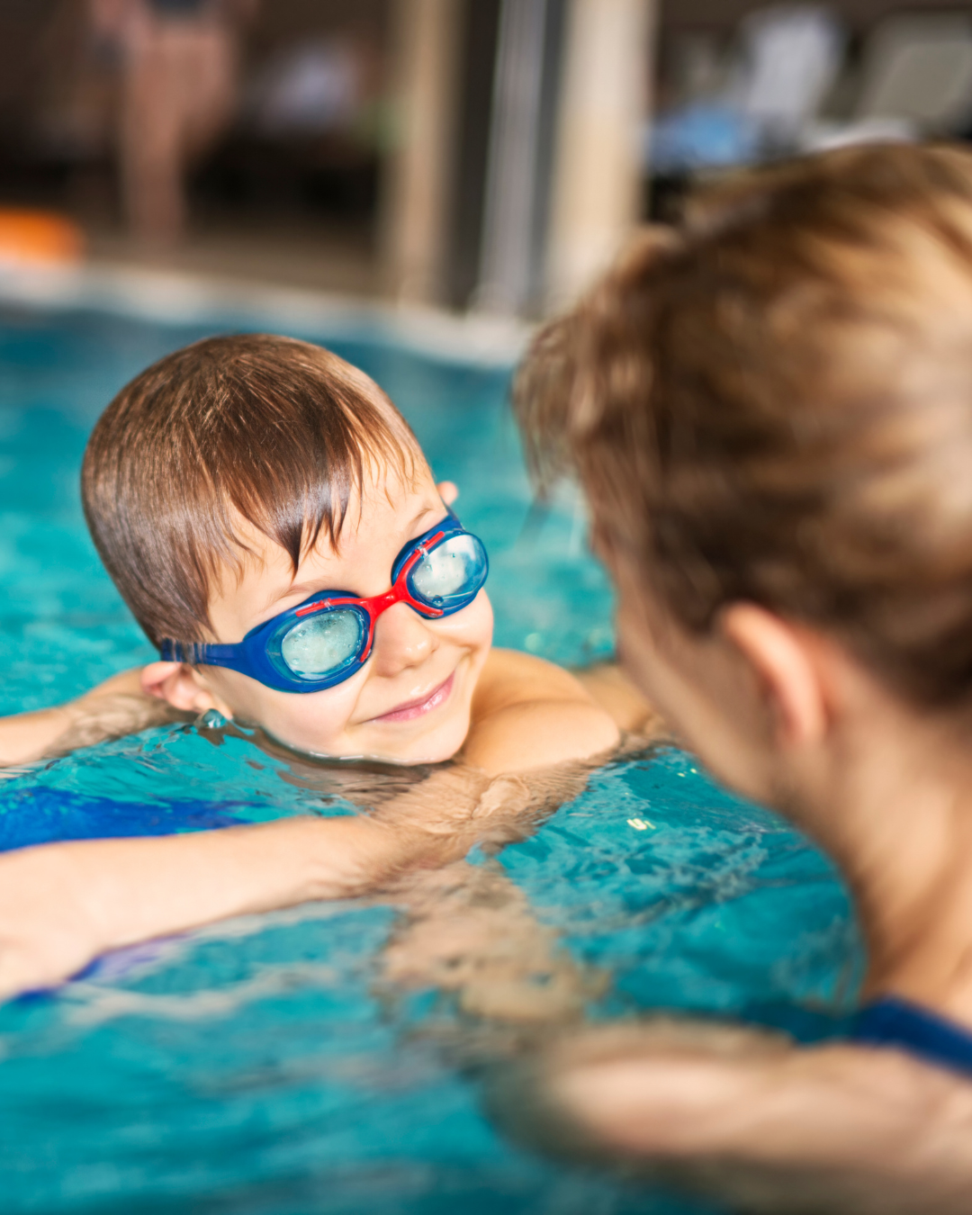 A young boy wearing blue and red swimming goggles smiling in a swimming pool, with an adult in the foreground.