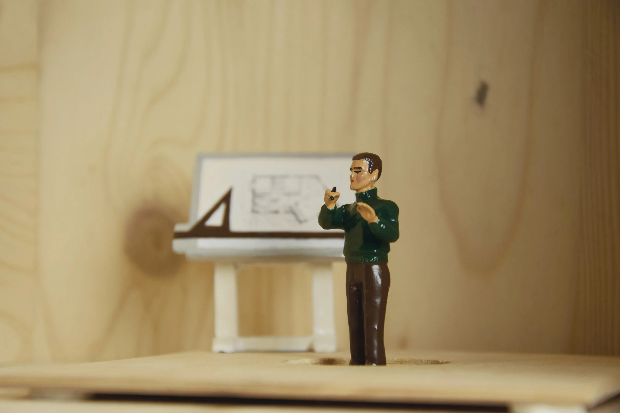 Miniature figurine of a man standing on a wooden shelf, holding a small object, with a small white table and framed picture behind him against a light wood-paneled wall.