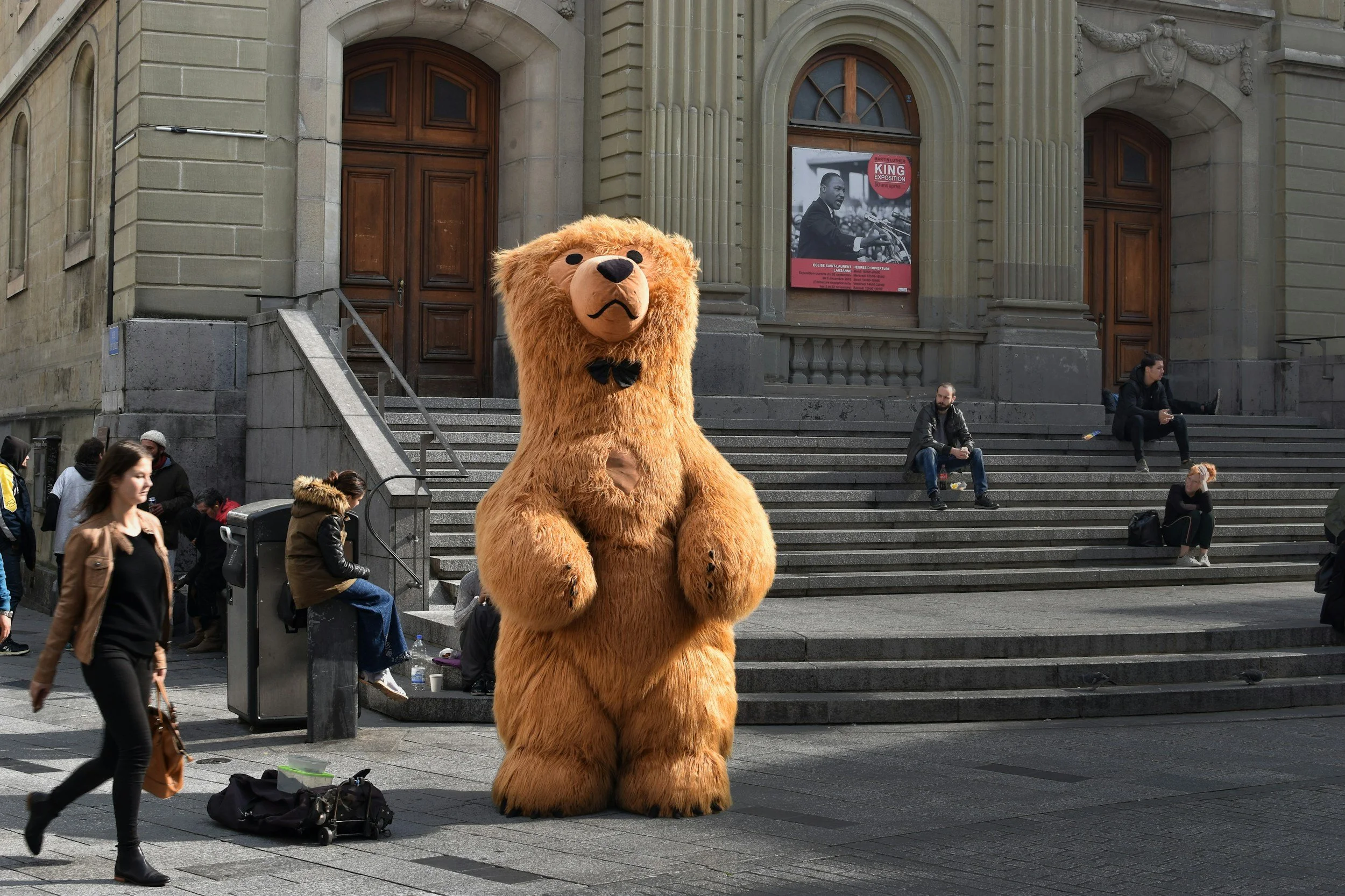 A person dressed in a large, furry lion costume standing in front of a stone staircase and a historic building entrance with wooden doors. People are sitting on the stairs and walking by in the background.