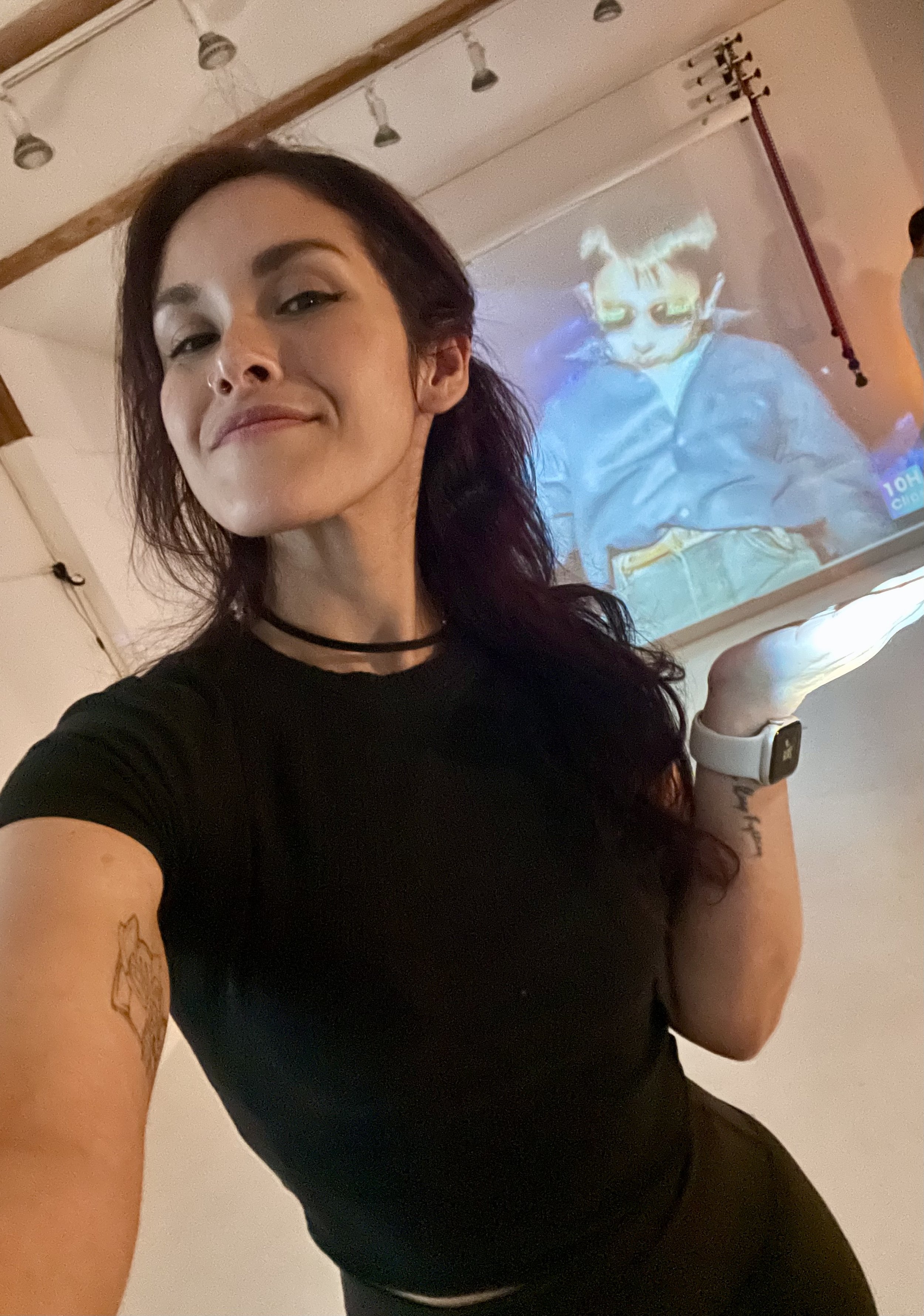 A woman with dark hair and a black shirt takes a selfie, showing her smiling face and tattoos on her left arm. Behind her, there is a projection of a cartoon character with animal features on the wall.
