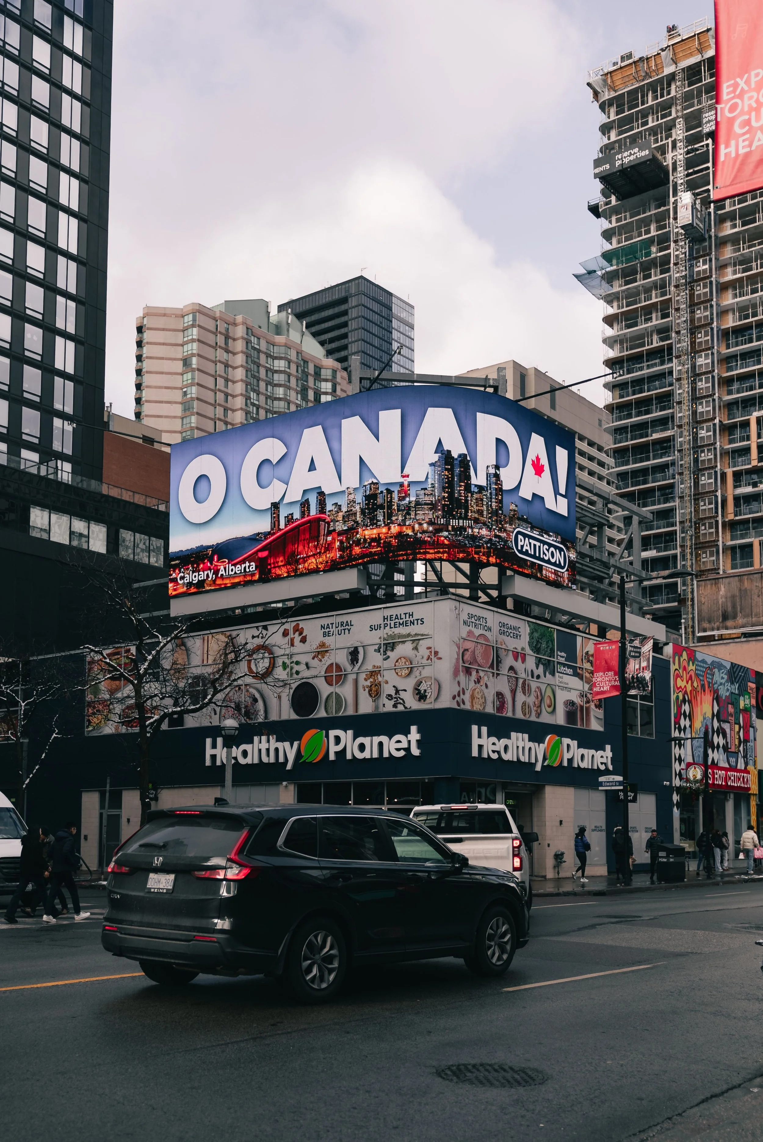 Cityscape with digital billboard displaying the message 'O CANADA!' with a view of tall buildings and a construction site. Cars are on the street with pedestrians walking on the sidewalk.