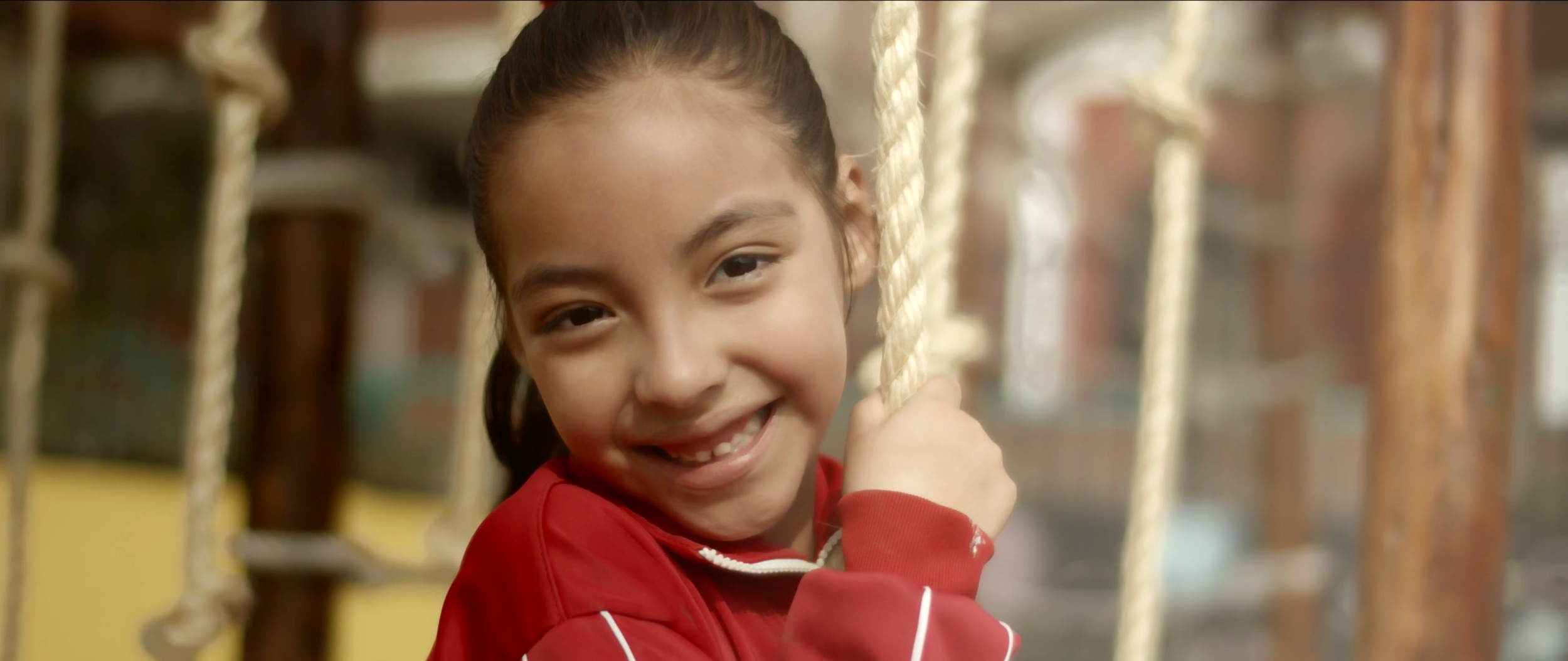 A young girl with dark hair in a ponytail smiling while holding onto a rope on a playground.