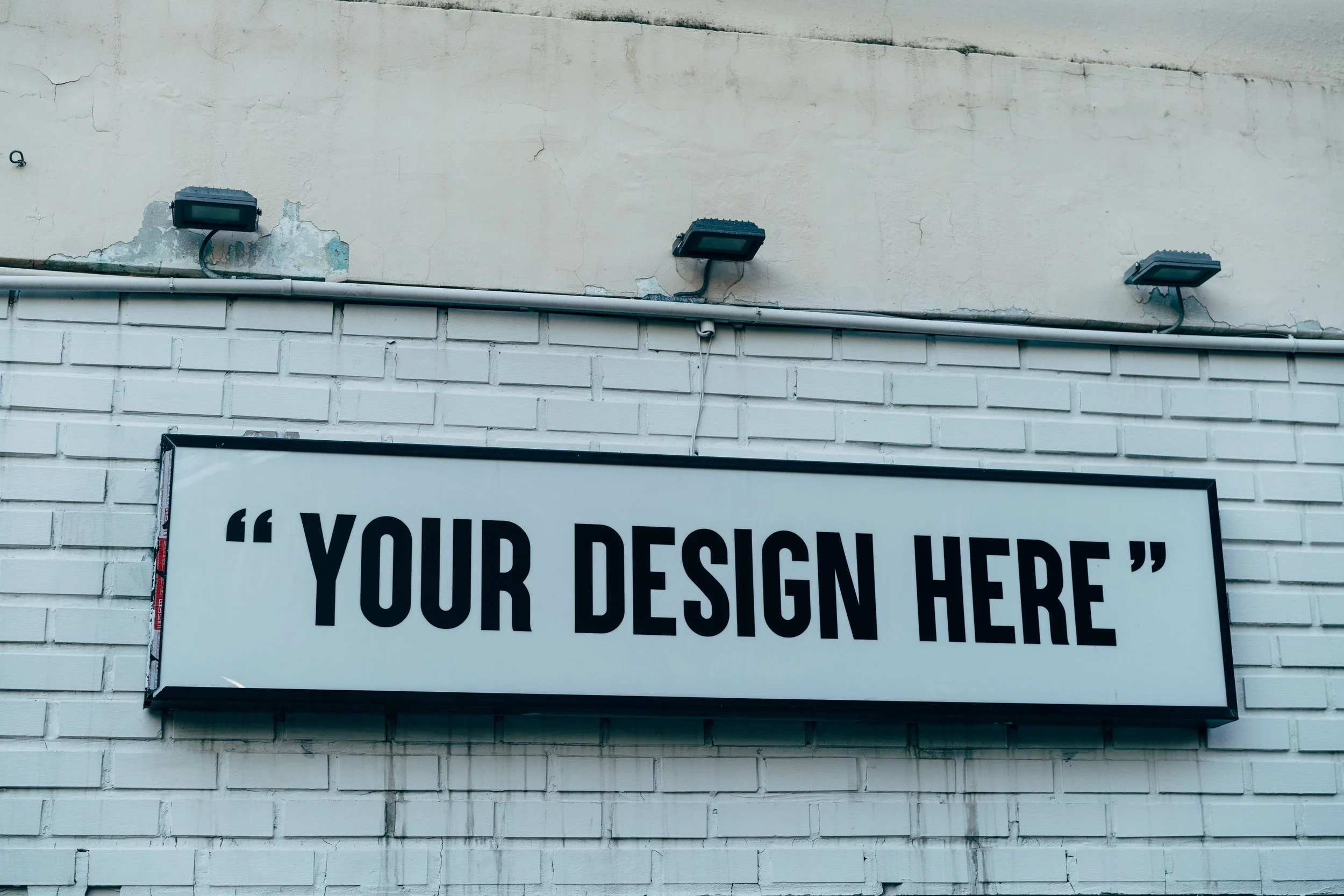 The image shows a white brick wall with three outdoor floodlights mounted above a framed board that has the words "YOUR DESIGN HERE" written in large black letters.