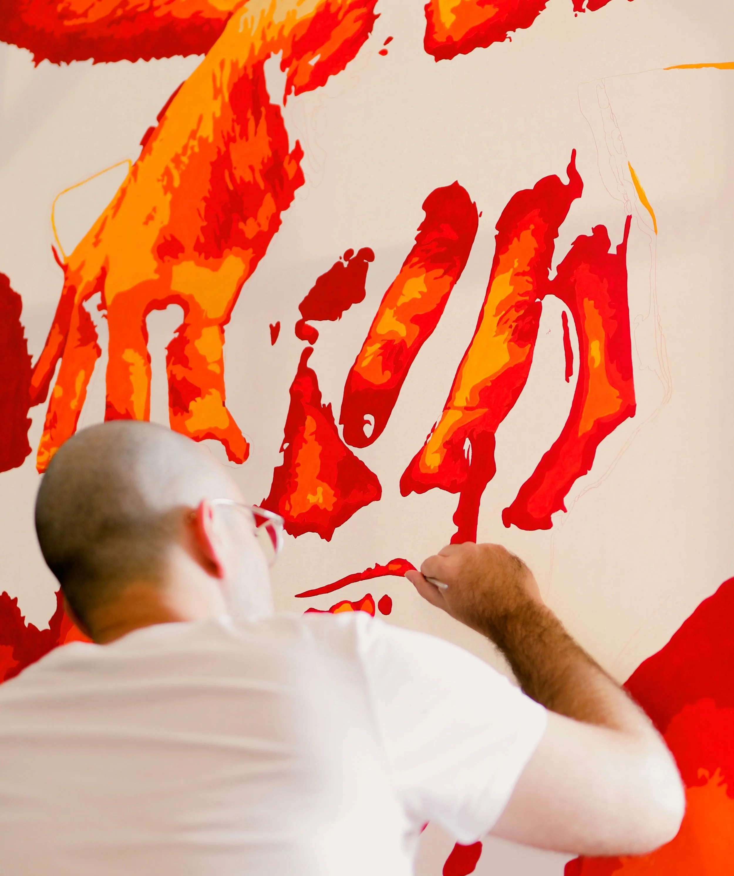 A person with a shaved head and white shirt painting an abstract artwork on a white wall using red, orange, and yellow colors.