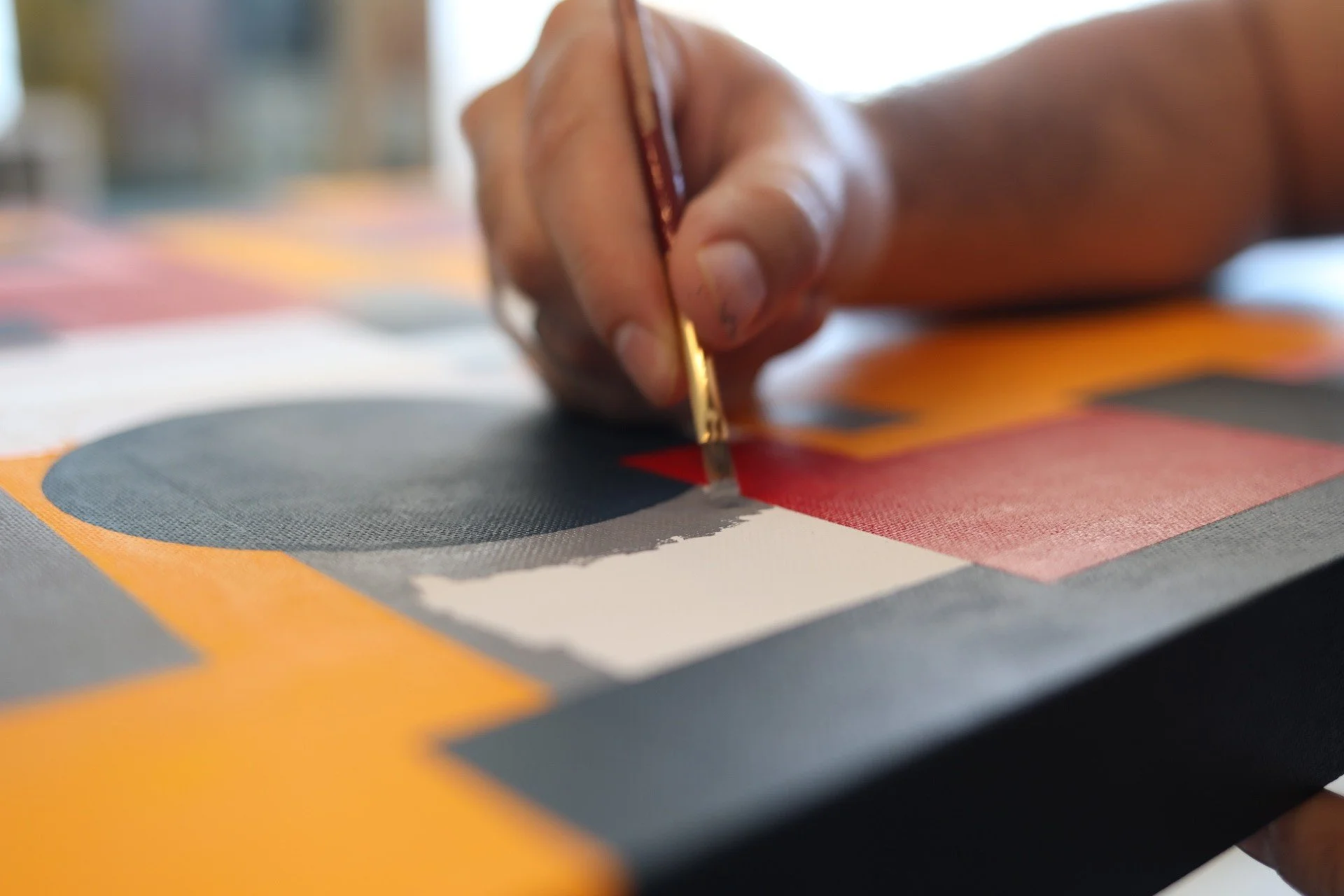 Close-up of a hand holding a paintbrush, painting geometric shapes in red, black, yellow, and gray on a canvas.