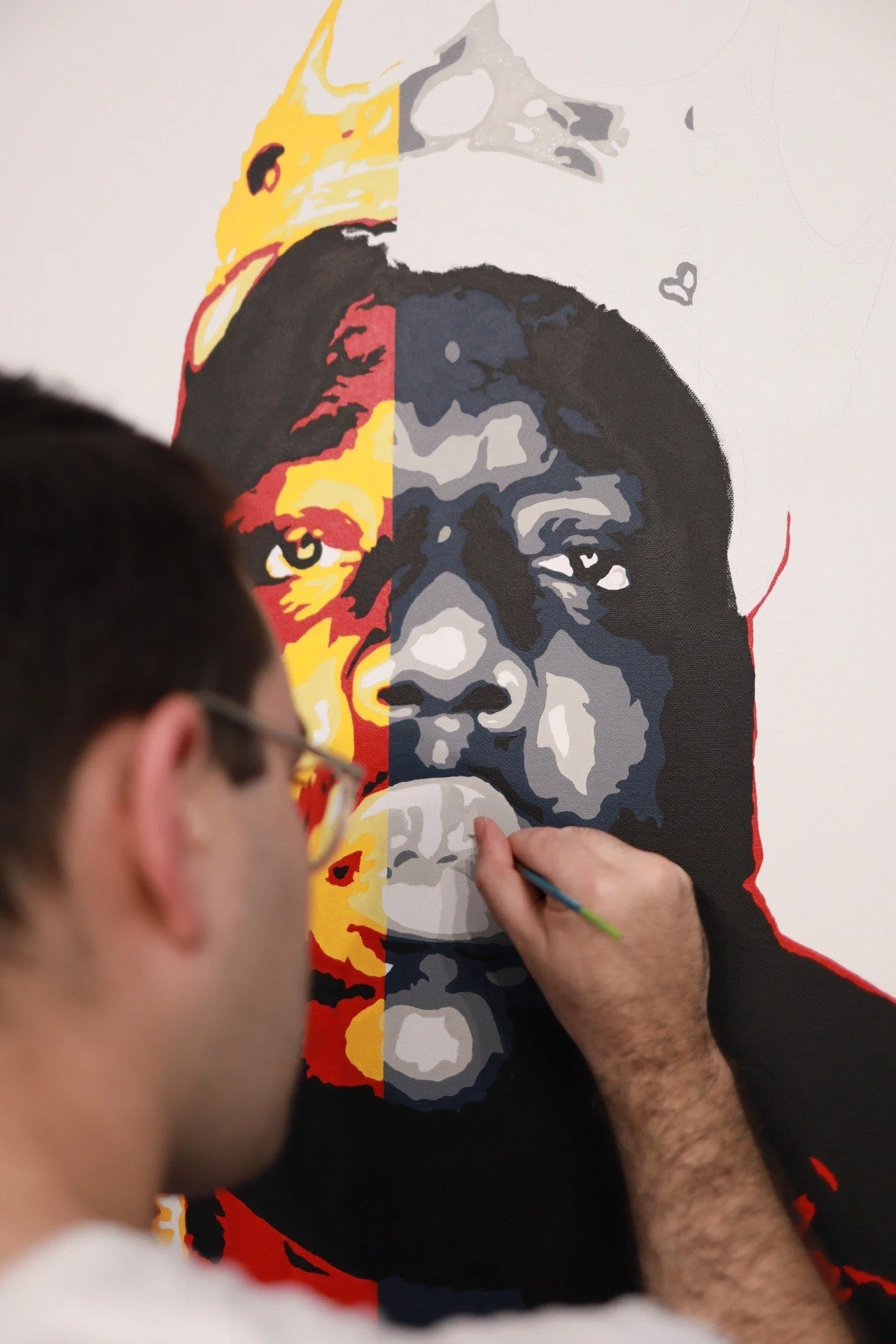 An artist is painting a large, colorful, portrait of a person's face on a wall. The face has different shades of black, gray, yellow, and red, creating a stylized artistic representation.