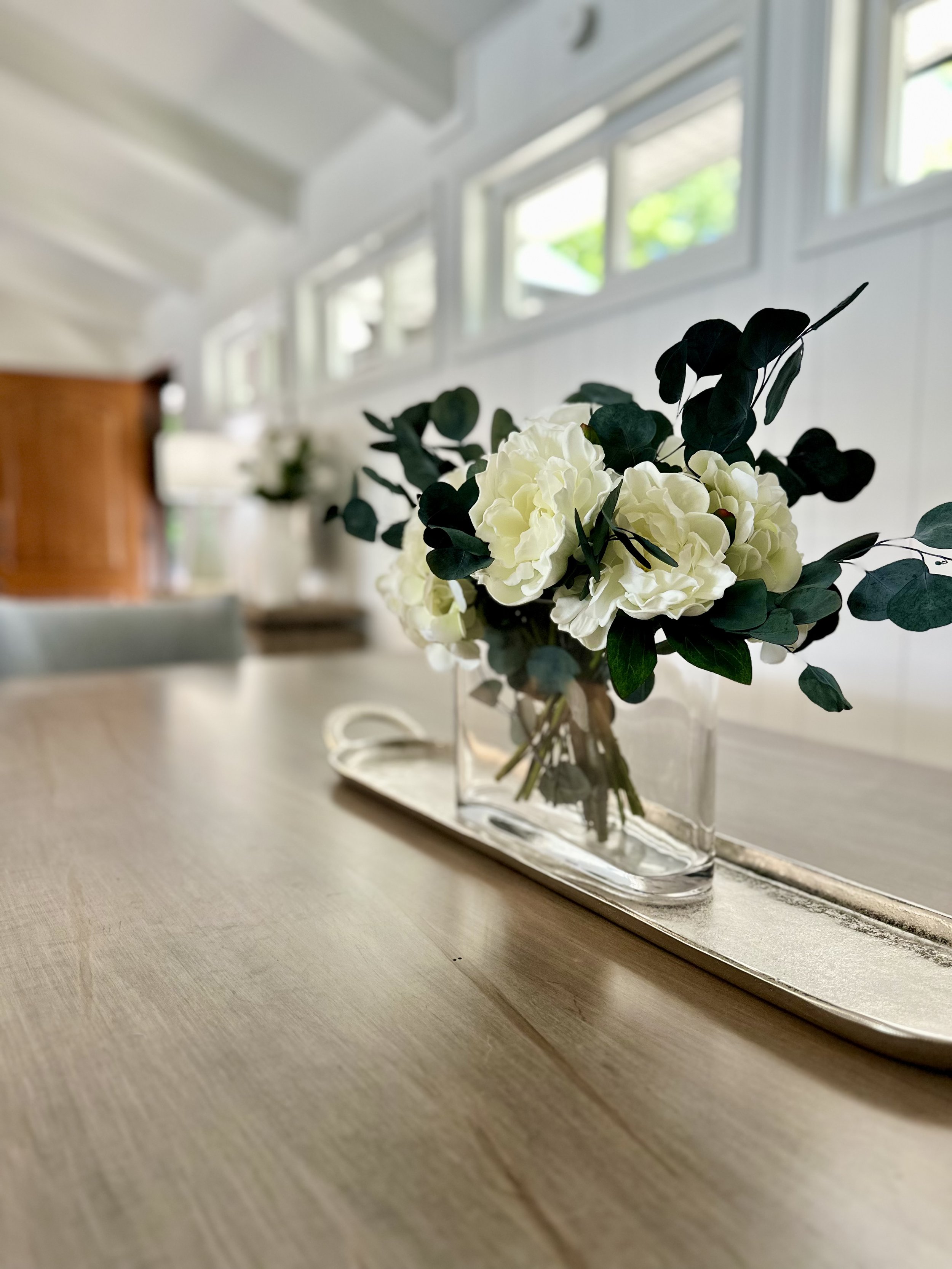 A clear glass vase with white flowers and green leaves on a wooden surface in front of a mirror, with windows and a white wall in the background.