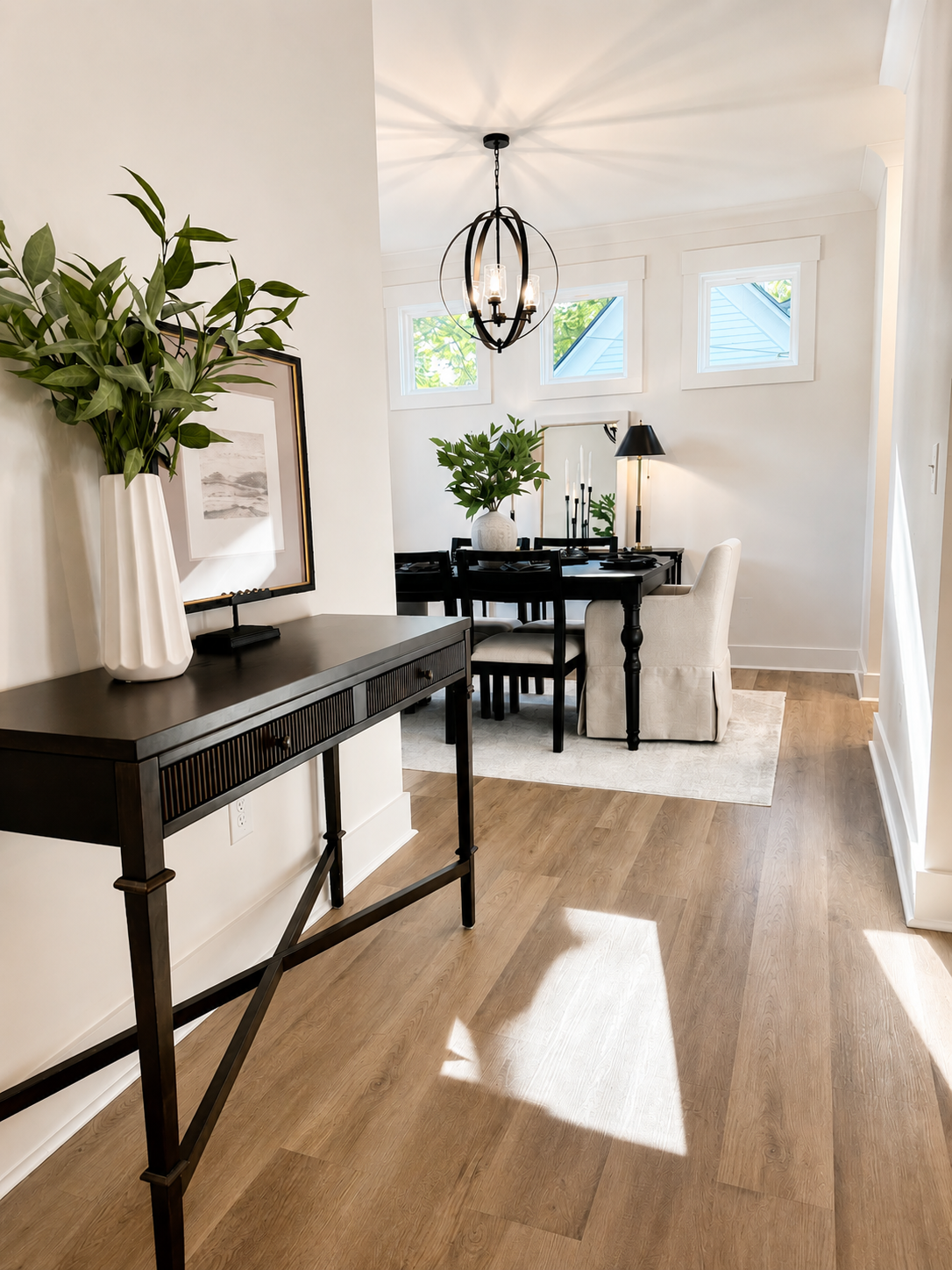 Dining area with black dining table, white upholstered armchair, black chairs, greenery, and modern lighting, in a bright room with large windows and wooden flooring.