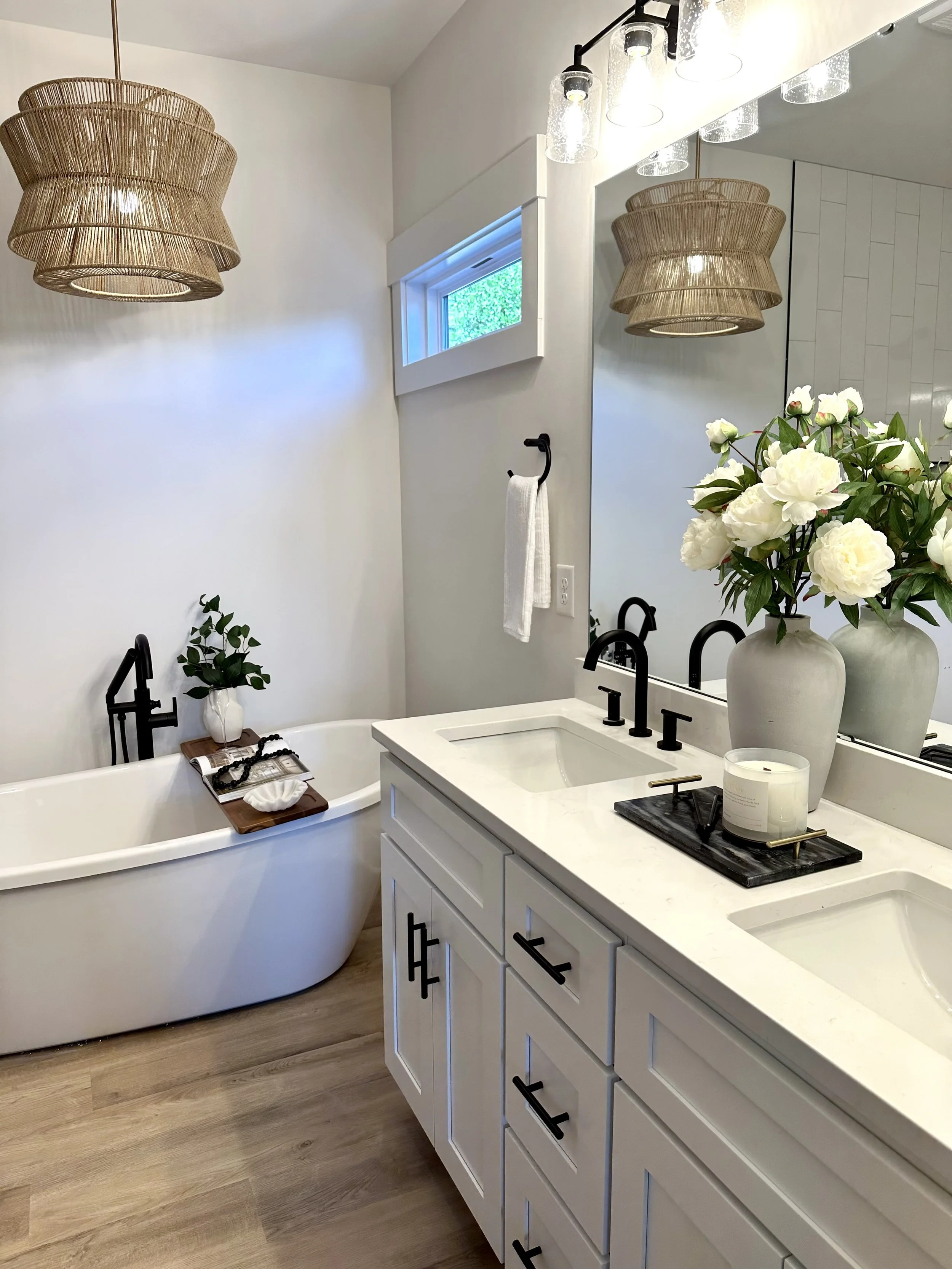 A modern bathroom featuring a white vanity with black hardware, a large mirror, a vase of white flowers, a black tray with a candle, a bathtub with a wooden tray holding books and a rolled towel, hanging wicker pendant lights, a small horizontal wind