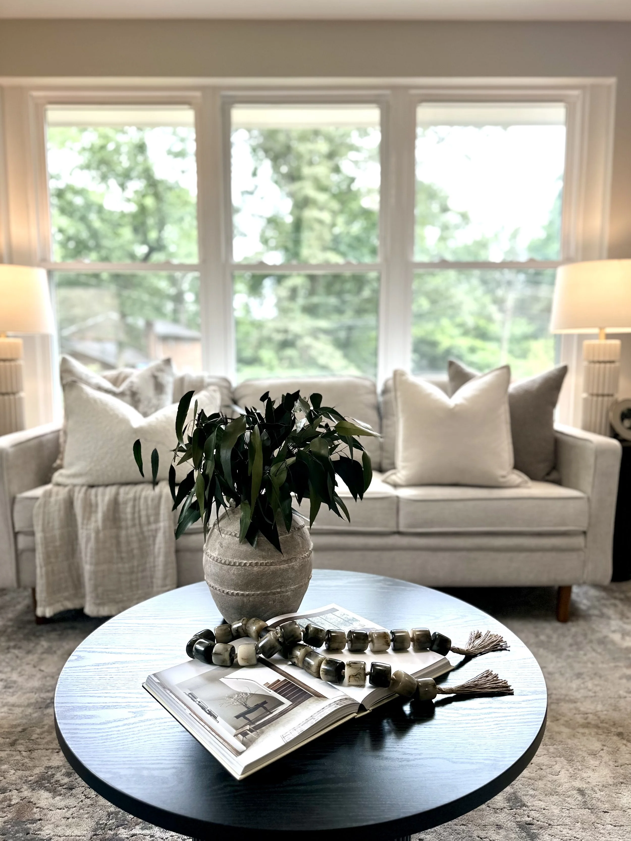 Living room with a beige sofa and pillows, large windows with trees outside, a round black coffee table with an open magazine, a beige vase with green leaves, a string of large beads, and two lamps on either side.
