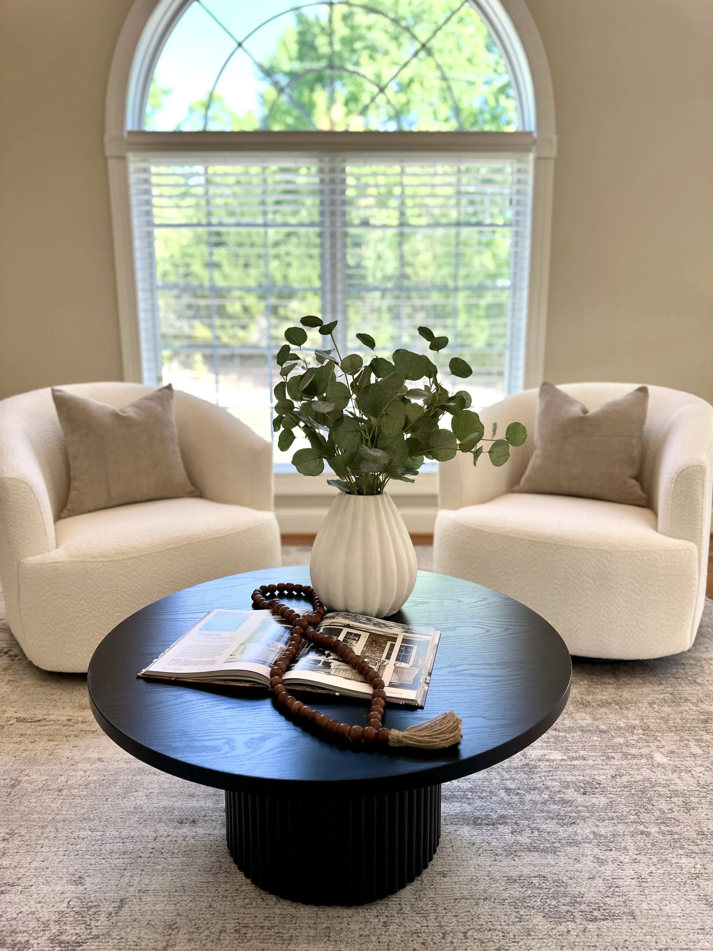 A living room with two white armchairs and beige pillows, a black round coffee table with an open magazine, wooden prayer beads, and a white vase with green foliage, placed in front of a large window with white blinds and a view of green trees outside.