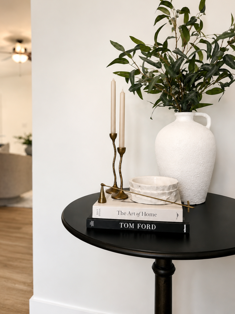 Decorative black round table with white textured vase holding green leafy plant, two beige candles in brass candlesticks, a white bowl, and two stacked books titled 'The Art of Home' and 'Tom Ford', in a bright room.
