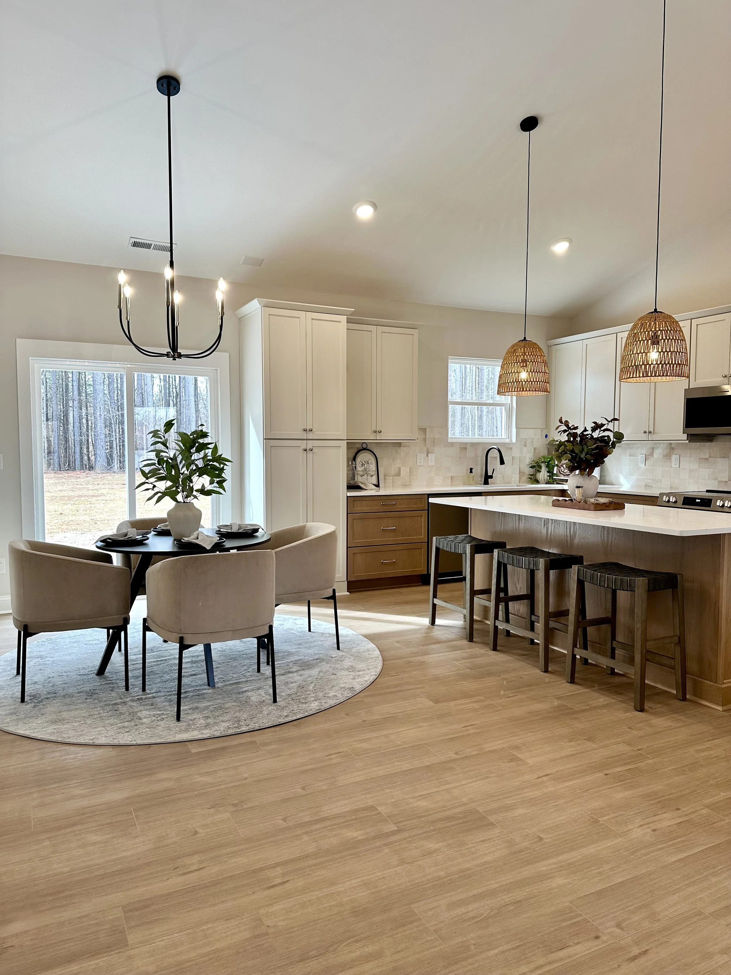 Modern kitchen with white cabinets, a kitchen island, two pendant lights, a dining table with four beige chairs, a vase with green plants, and large windows overlooking a wooded yard.