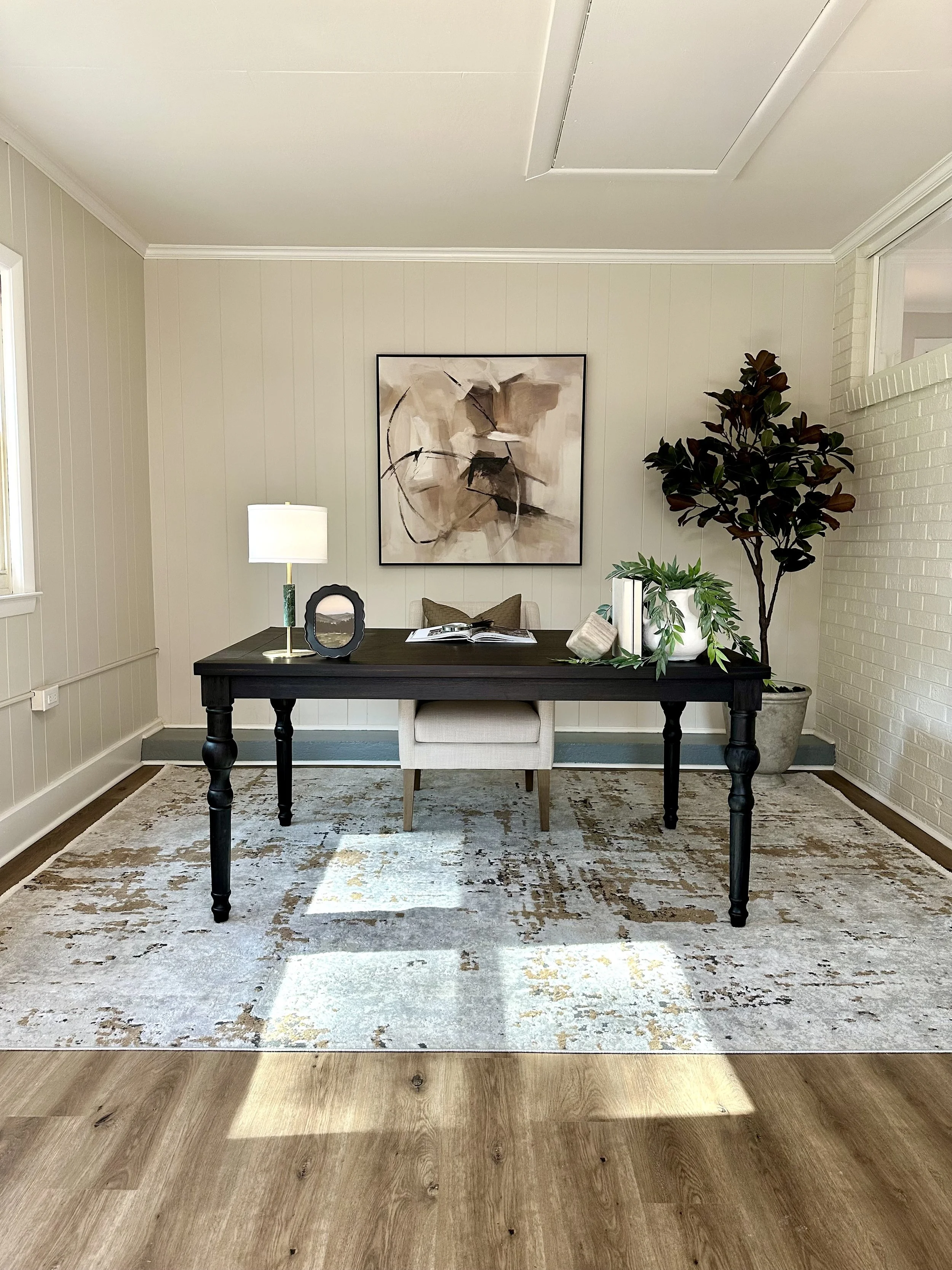 A dining room with a black wooden table, a white upholstered chair, a beige and gray abstract painting on the wall, a white table lamp, and various decorative items and plants on the table. The room has light-colored paneled walls, a beige and gray p
