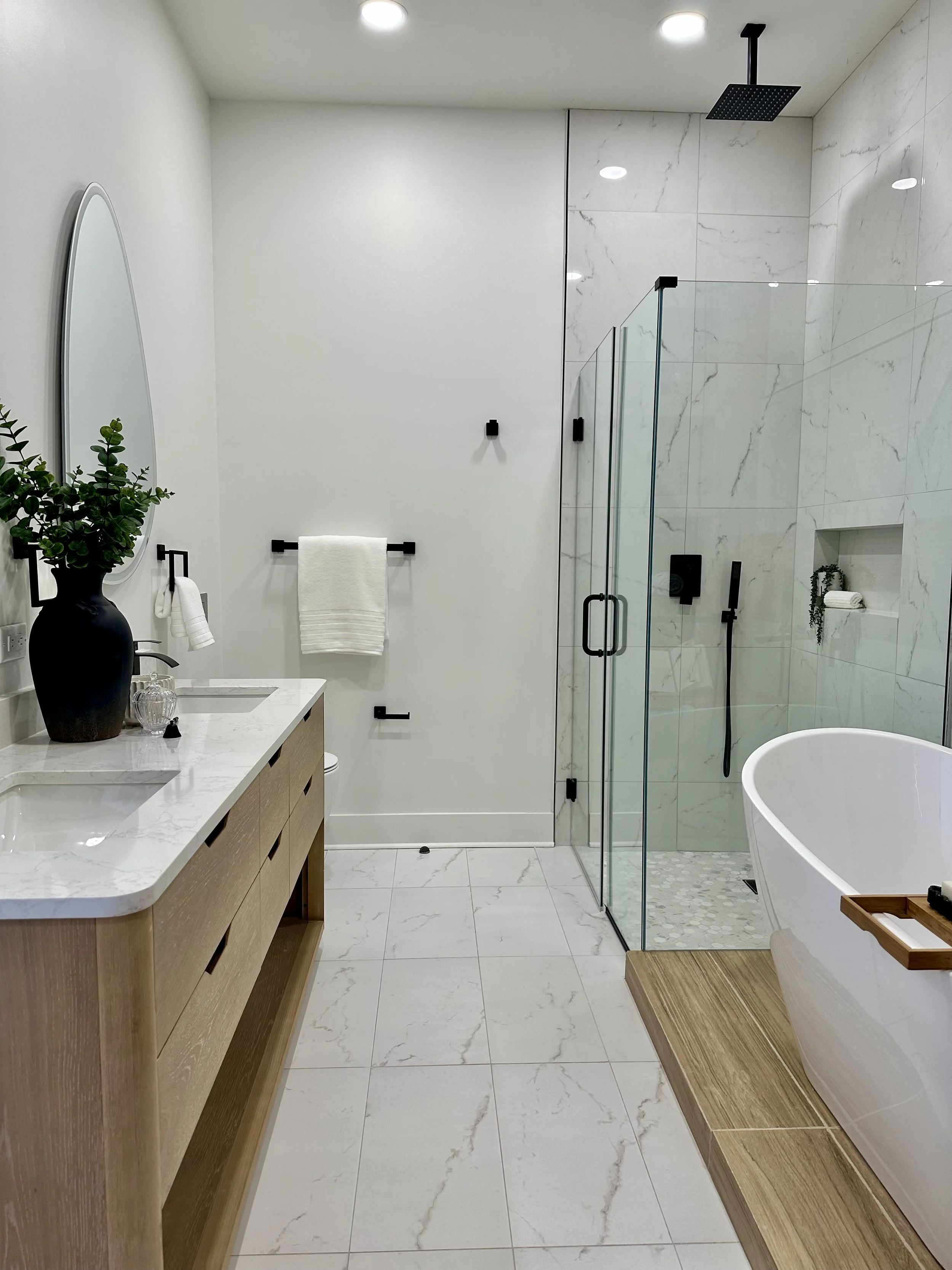 Modern bathroom featuring a freestanding bathtub, a walk-in shower with glass doors, a wooden vanity with a white marble countertop, a large mirror, a potted plant, white tiled floors and walls, and black fixtures.