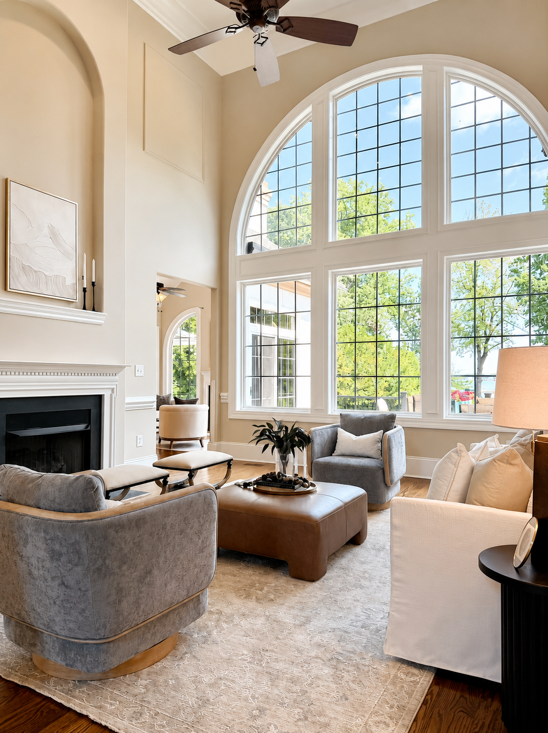 Bright living room with large arched windows, beige walls, and a sitting area including a gray armchair, a white sofa, a brown leather ottoman, and a beige rug. There are decorative candles on the mantel, a potted plant, and a table lamp.