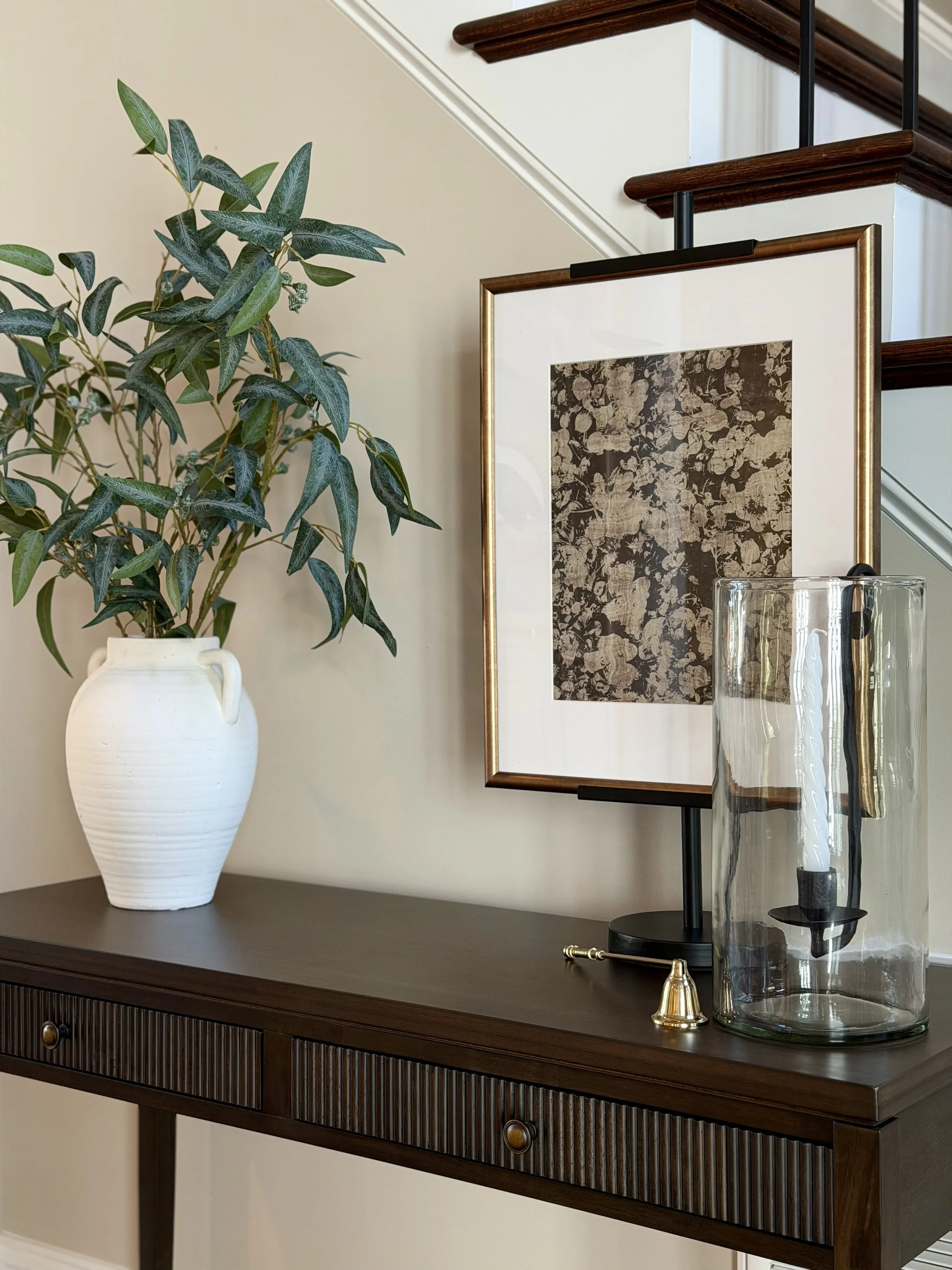 A dark wood console table with a curved front, ribbed drawer panel, and round brass knobs. On top, a white ceramic vase with green leafy plant, a framed abstract floral artwork, and a tall cylindrical glass vase with black decorative objects inside, 