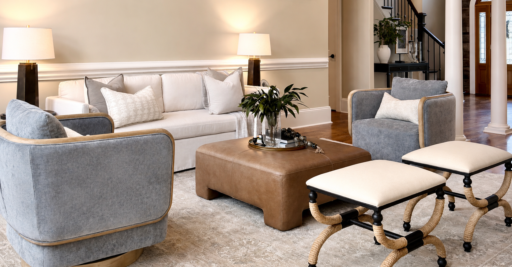 A living room with a white sofa, gray armchairs, a brown leather ottoman with a tray of decor including a plant, a candlestick, and books, two lamps on black side tables, and additional chairs with curved wicker legs and white cushions. There is a staircase in the background and natural light coming through the front door.