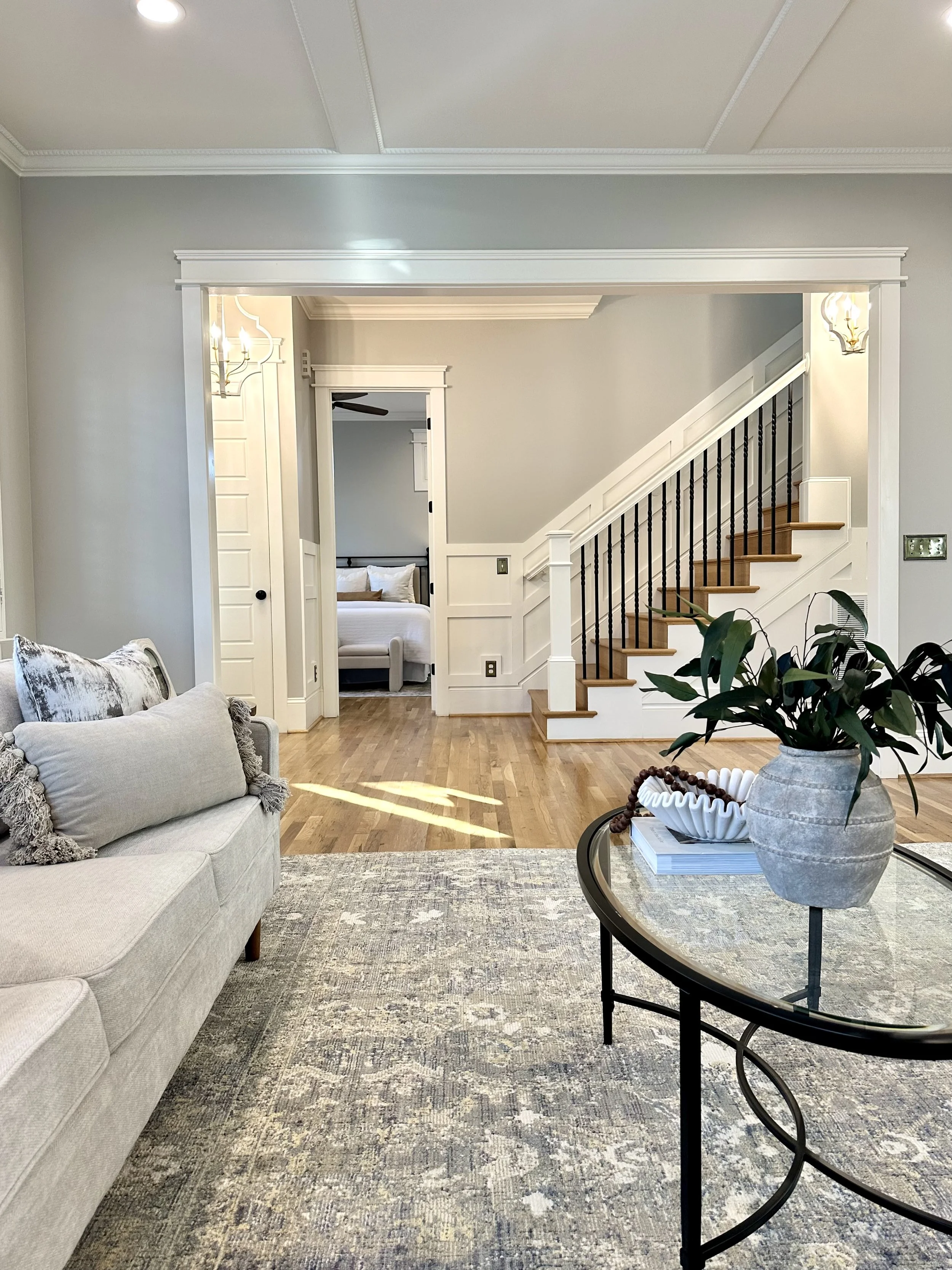 Living room with light-colored sofa and decorative pillows, patterned area rug, round glass-top side table with potted plant, staircase with black balusters and white railing, doorway to a bedroom with bed, crown molding and wall-mounted light fixtur