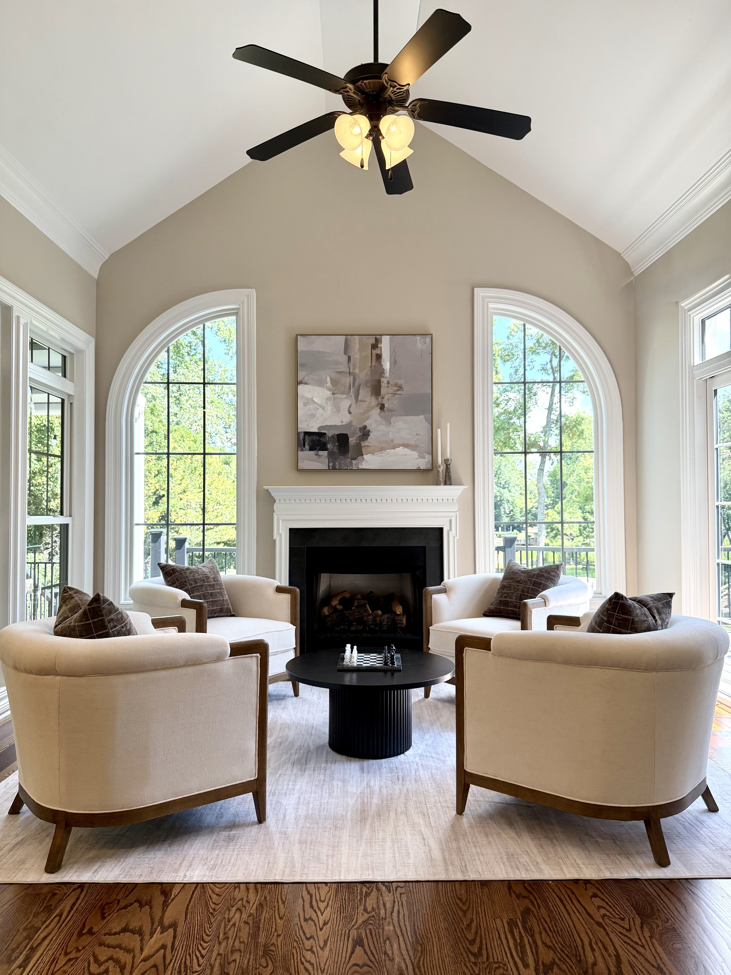 Living room with large arched windows, a white fireplace, and four beige armchairs with brown pillows arranged around a black round coffee table with a chessboard on it. There is a ceiling fan with lights and a modern abstract painting above the fire