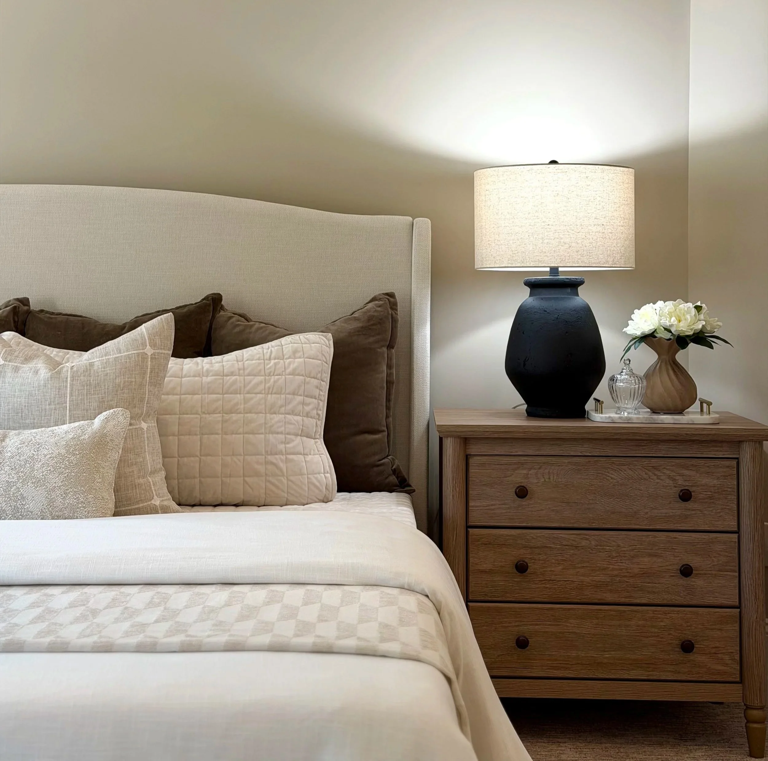 A bedroom scene with a white bedspread, beige and white throw pillows, a brown plush blanket, a light wood nightstand, a black table lamp with an off-white shade, a small bouquet of white flowers in a glass and ceramic vase, and a beige wall with a white upholstered headboard.