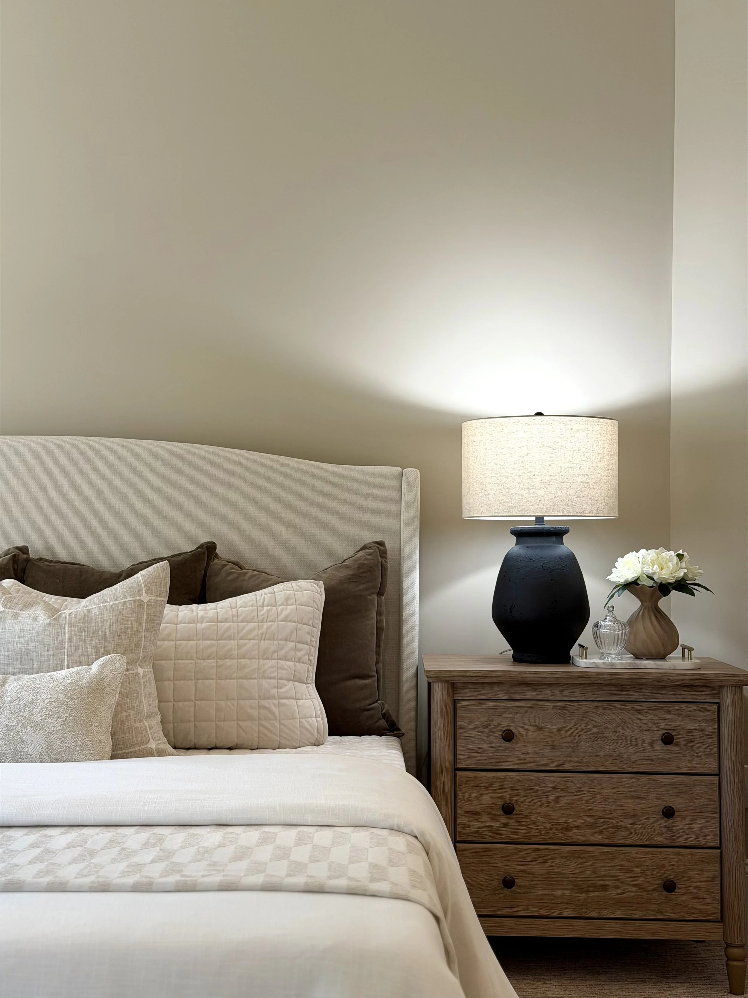 A bedroom corner with a bed, a beige headboard, multiple pillows, a wooden bedside table, a black vase with white flowers, a beige table lamp, and a neutral wall.