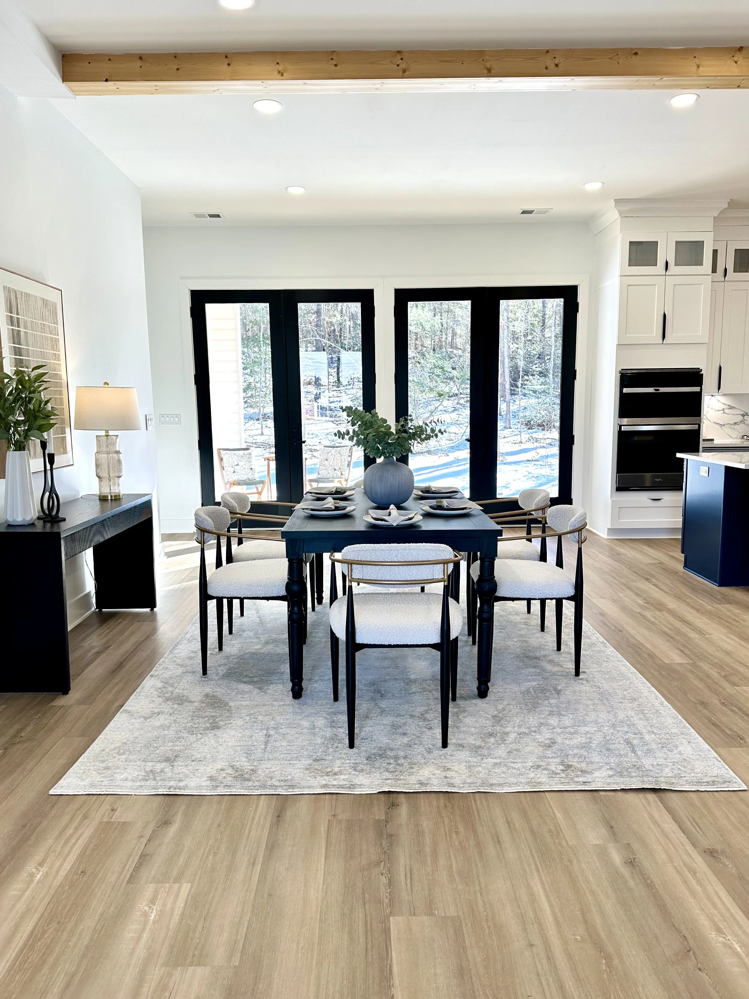 Dining room with a dark wooden table, six light-colored upholstered chairs, a large gray rug, a decorative vase with greenery, white cabinetry, black appliances, and large windows with outdoor trees visible.