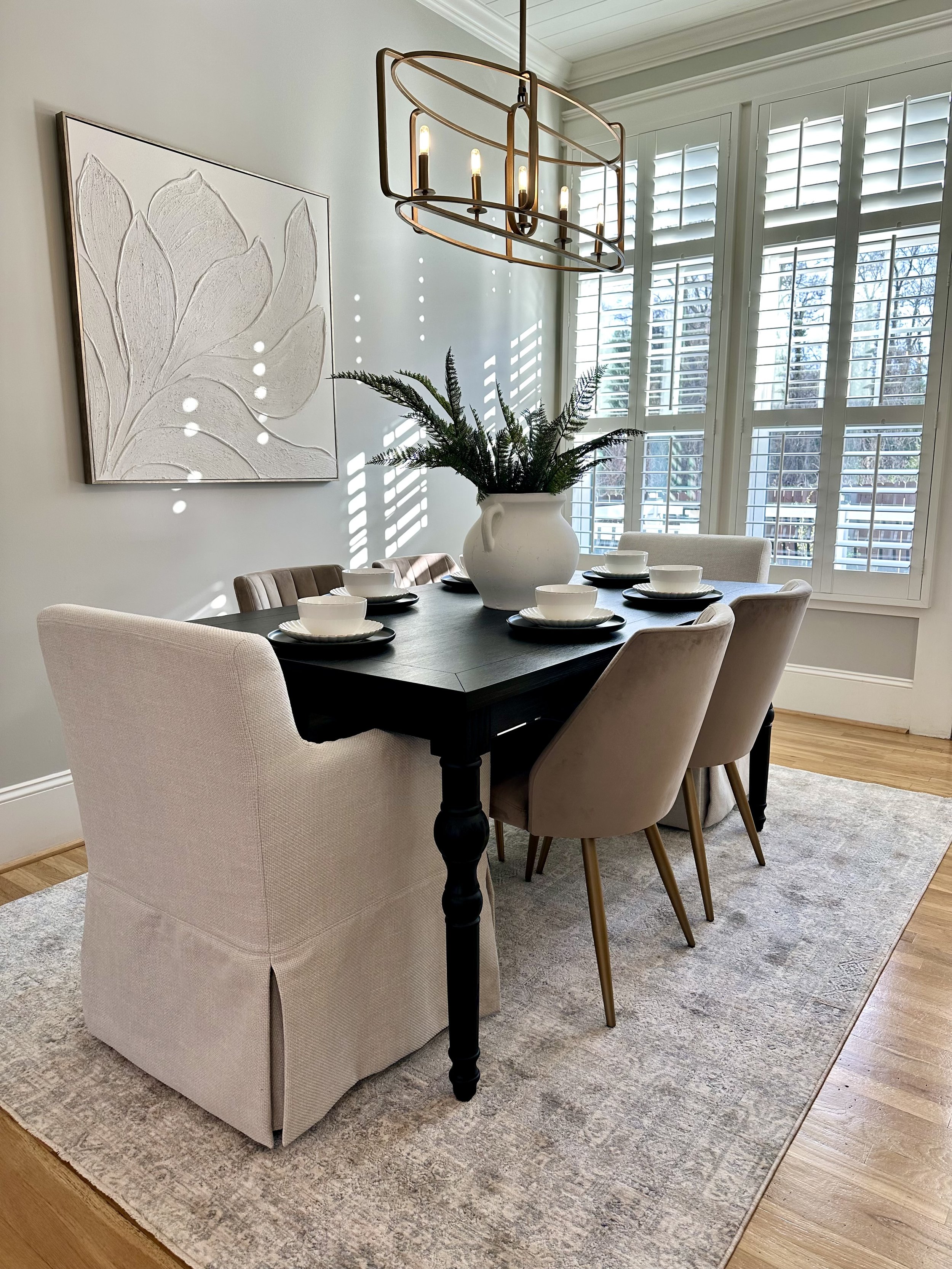 A dining room with a black table, beige chairs, a large white vase with greenery, wall art, a large window with shutters, and a modern chandelier hanging above the table.