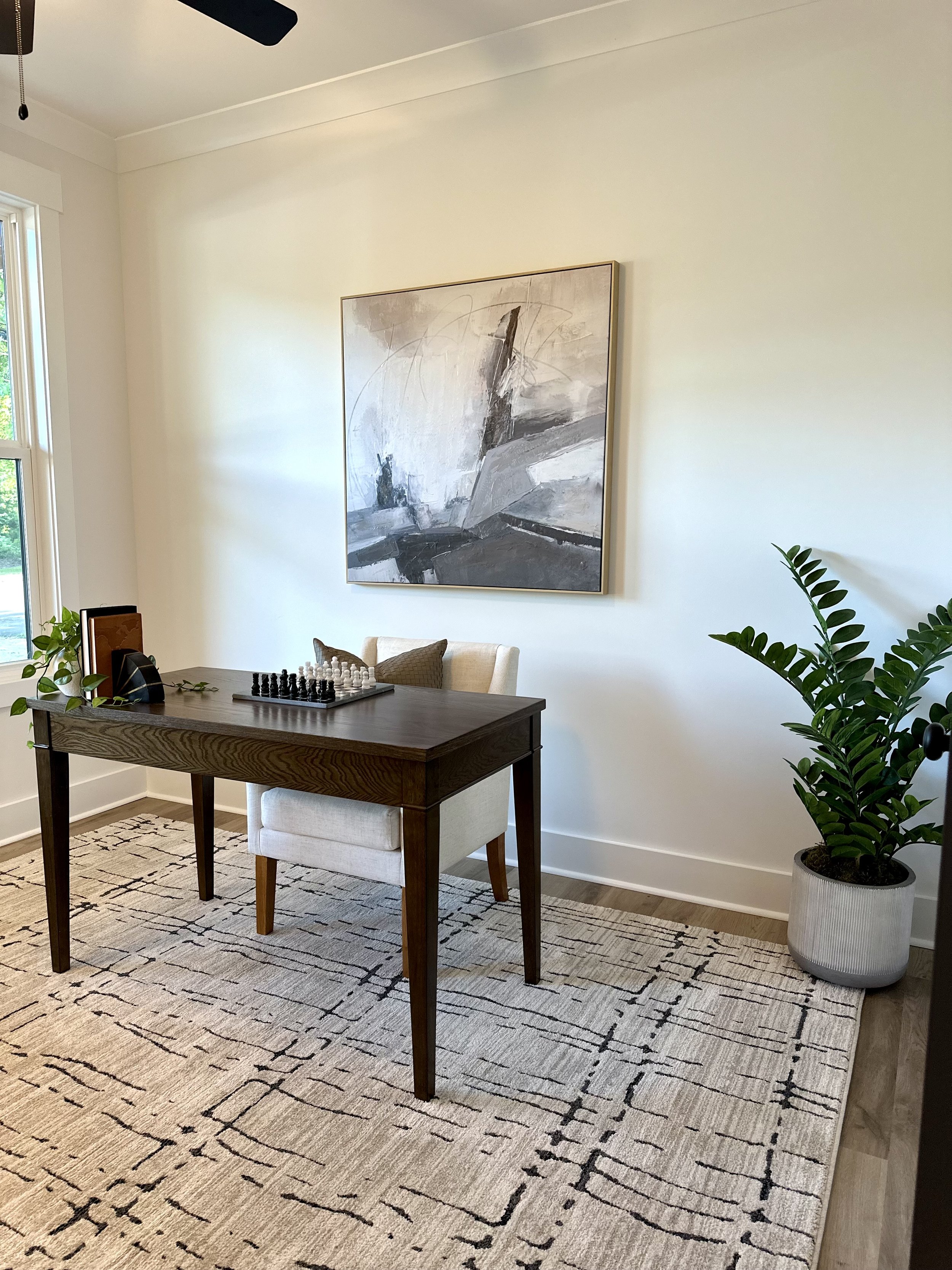 A room with a wooden dining table, a white upholstered chair, a chessboard with pieces, large abstract black-and-white artwork on the wall, a potted plant, and a light-colored patterned rug.