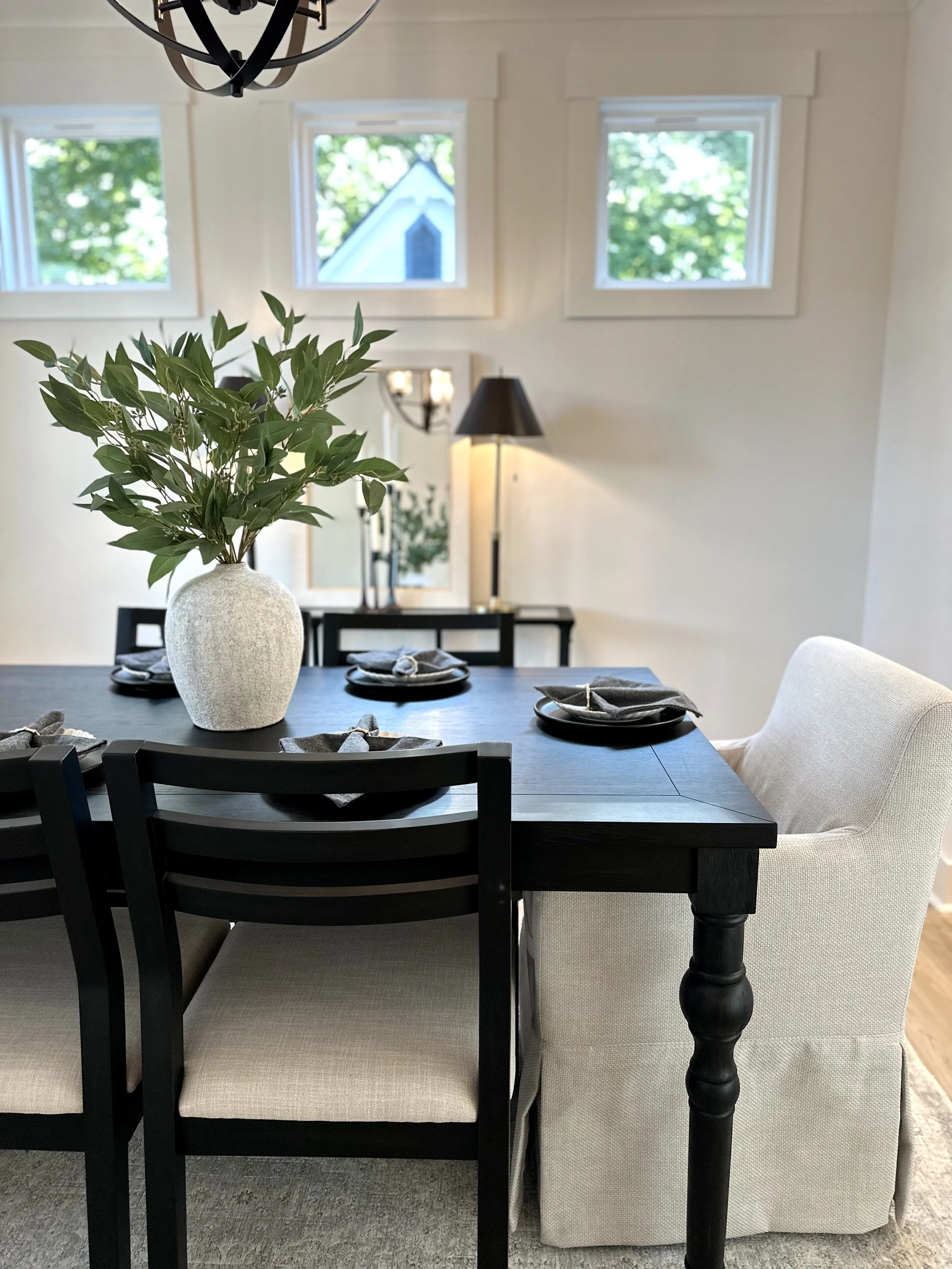 A dining room with a black table set with plates, napkins, and utensils, a large white textured vase with green leaves, and cream upholstered chairs. There are three small windows with greenery visible outside, a black floor lamp, a mirror, and a lar