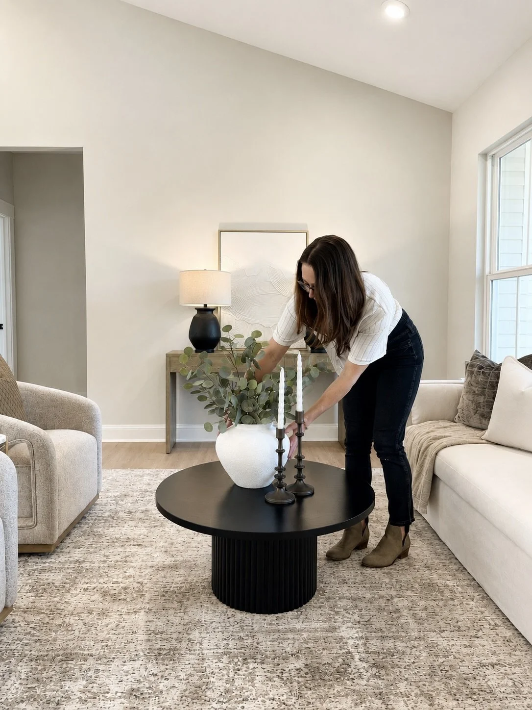 Woman arranging a plant on a black coffee table in a living room with beige furniture, a lamp, and decorative items.