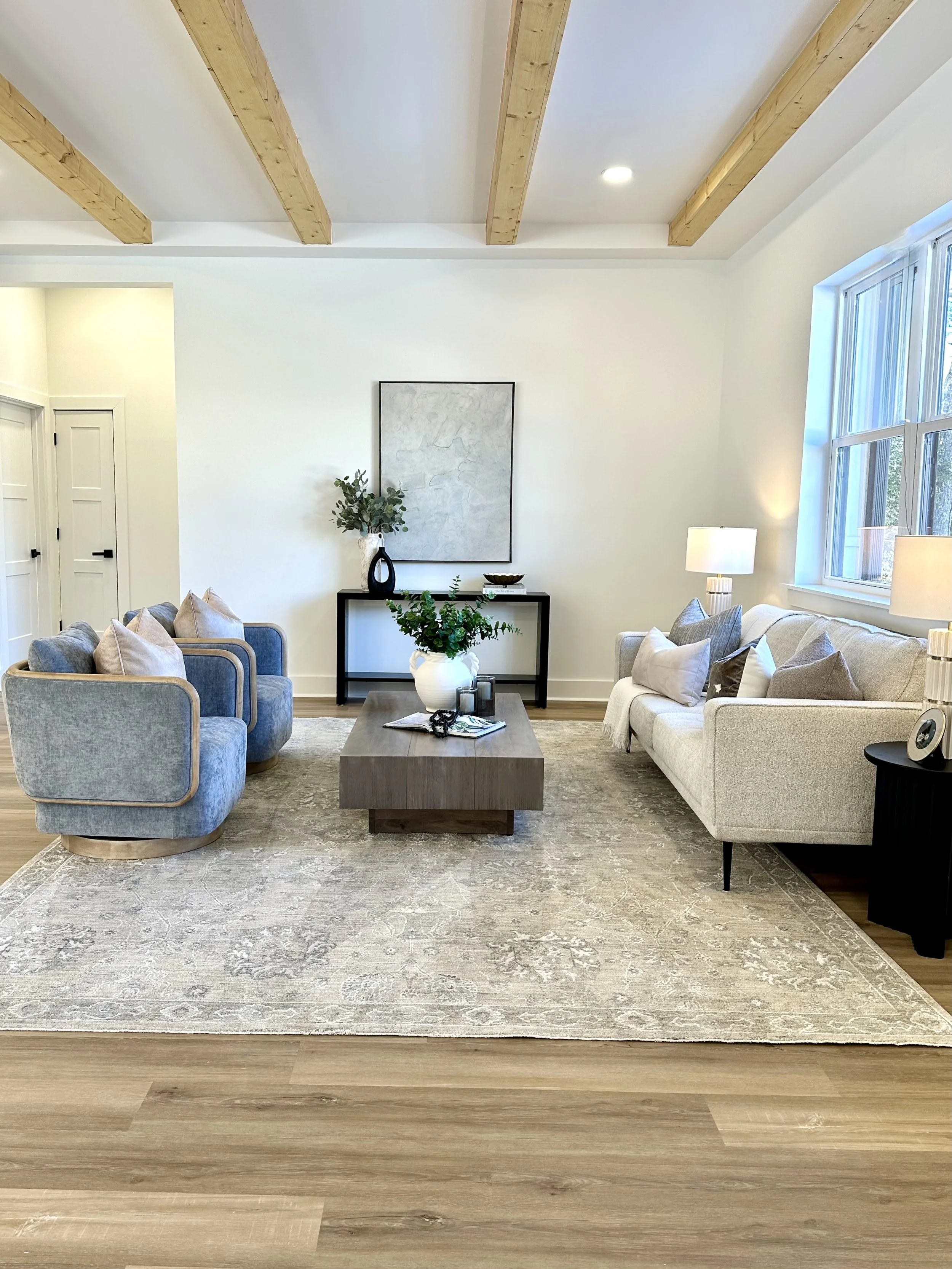 Living room with light-colored sofa and blue armchairs arranged around a wooden coffee table, with a black console table, artwork, and decor, large window, and a beige rug.