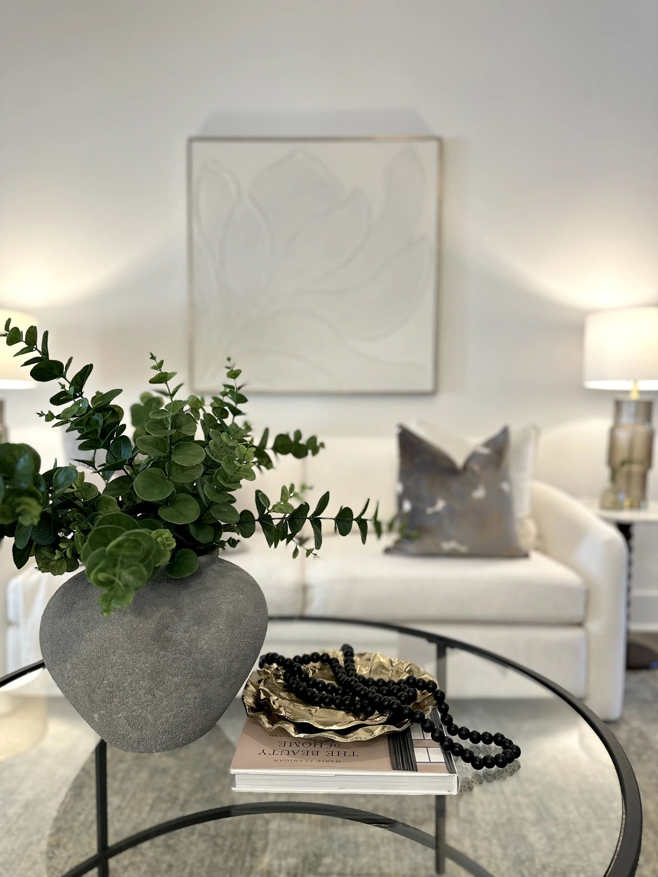 Close-up of a gray textured vase with greenery, on a black metal table with decorative objects including a gold dish with black beads and a magazine, in a living room with artwork and lamps.