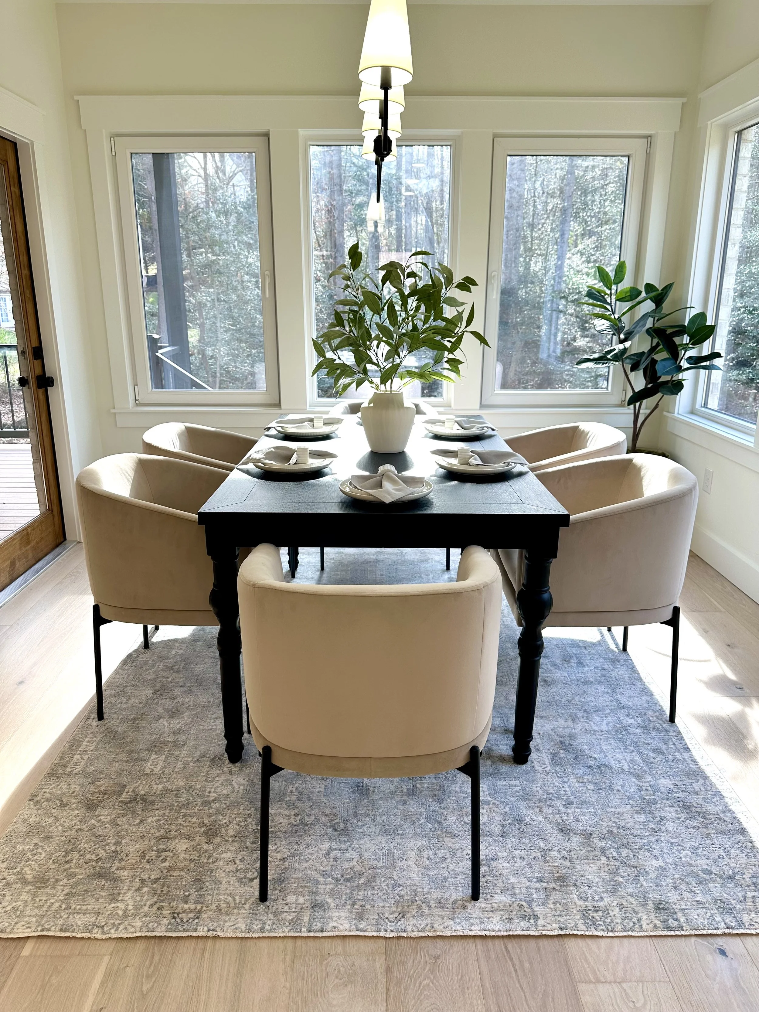 A dining room with a rectangular dark wood table set with white plates, napkins, and utensils for six people. Six light beige upholstered chairs surround the table. A large white ceramic vase with green leafy plants decorates the center of the table.