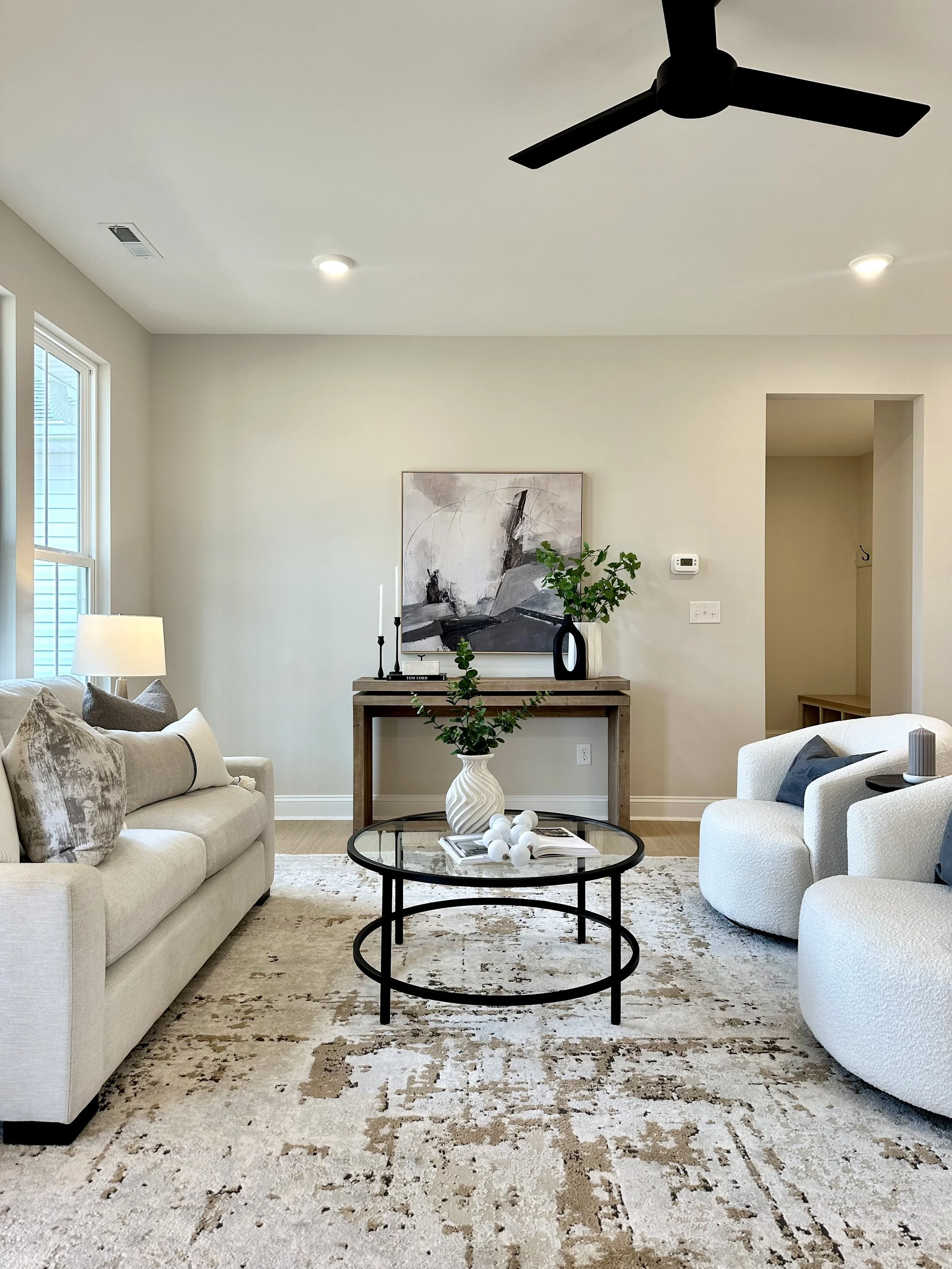 Living room with beige couch and armchair, black oval coffee table, abstract artwork on wall, green plant in vase, white rug, ceiling fan, window with blinds, and a side table with lamp.