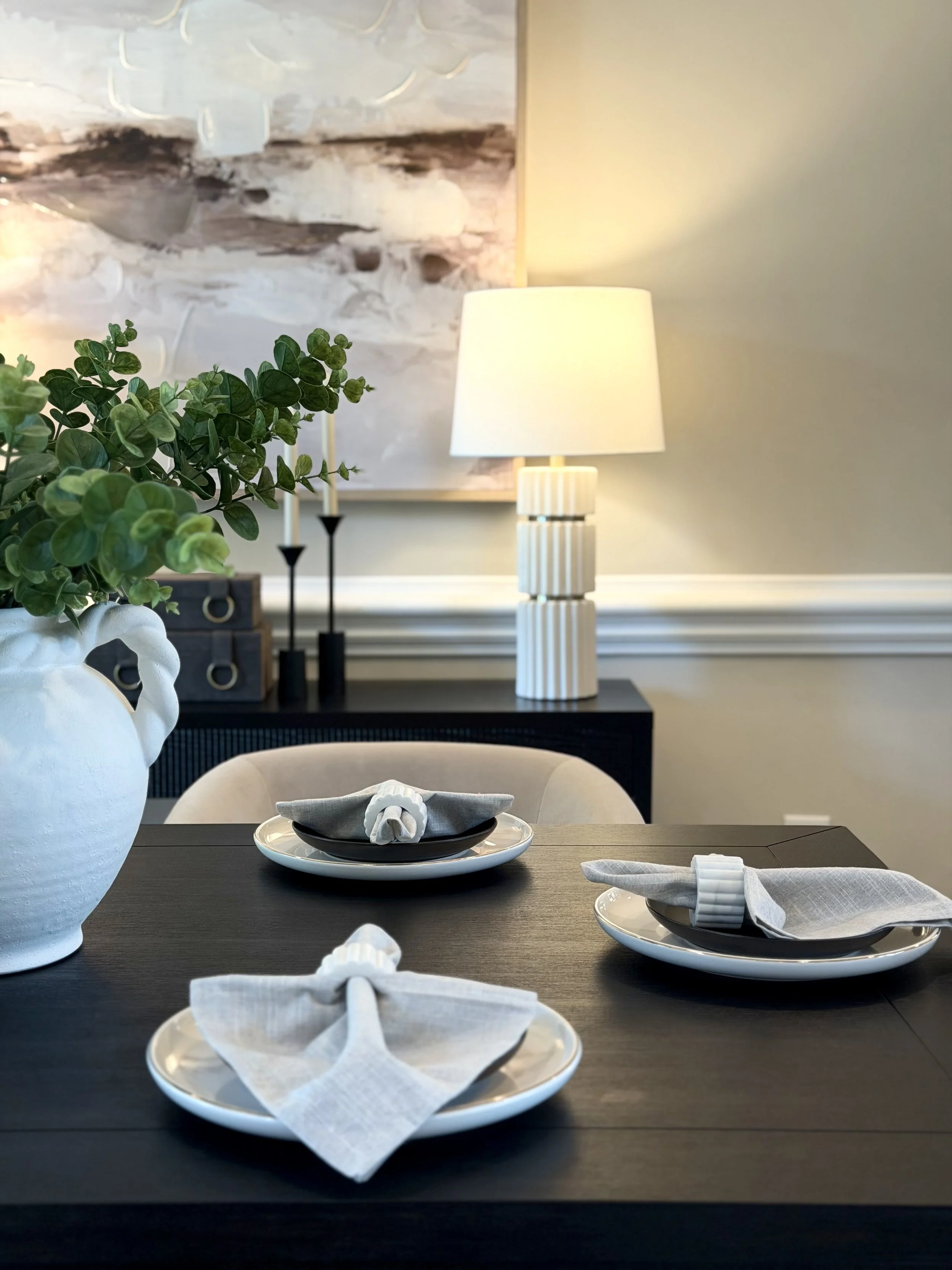 A dining table set with white plates, black bowls, gray napkins, and white linen napkins shaped like flowers. A large white vase with green foliage, a white lamp with a striped base, and decorative black candle holders are in the background. A painti