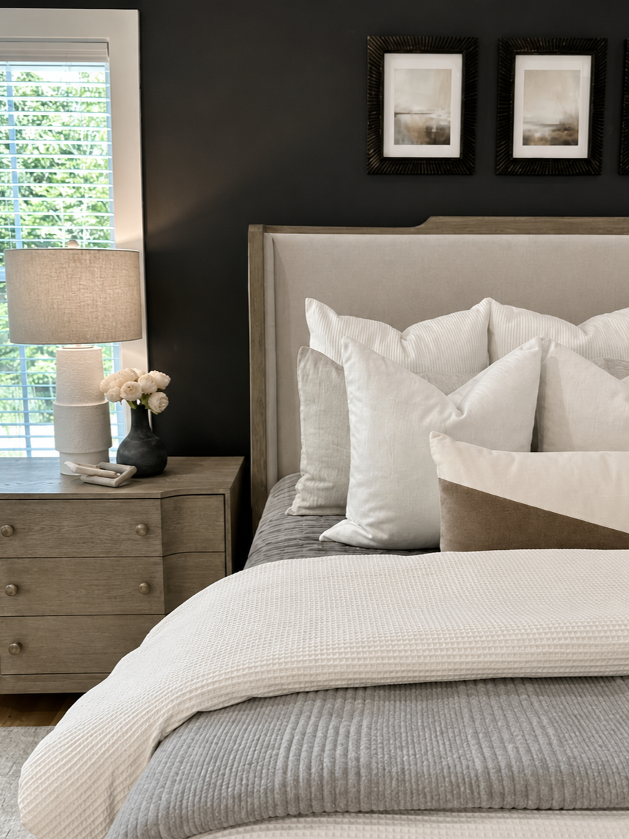 A neatly made bed with a beige upholstered headboard, white pillows, and a textured white blanket, next to a wooden nightstand with a table lamp, a vase of white flowers, and a window with blinds, in a bedroom with dark gray walls and framed artwork.