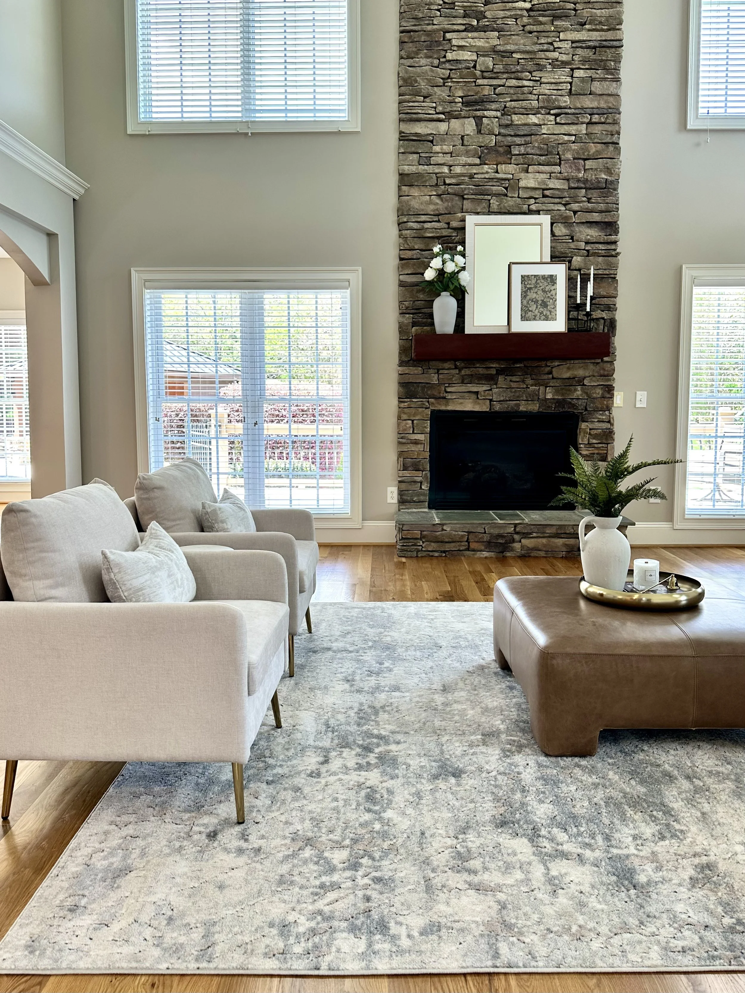 Living room with beige sofa, light blue patterned rug, brick fireplace with decorative items on mantel, large windows with blinds, and wooden flooring.