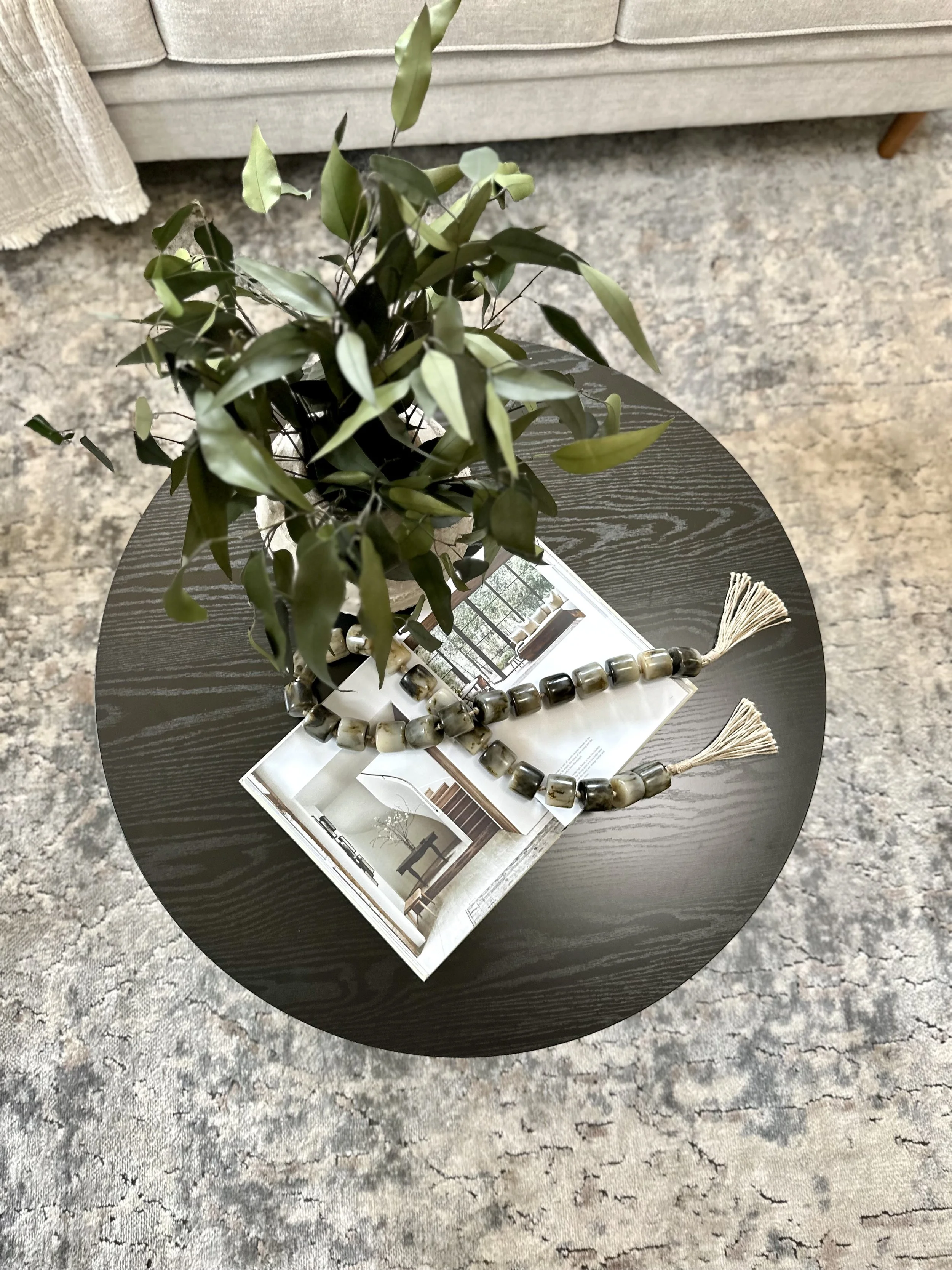 A round black table with a potted green plant, an open magazine showing interior room design, and a beaded necklace with tassels, on a mottled gray and tan carpet.