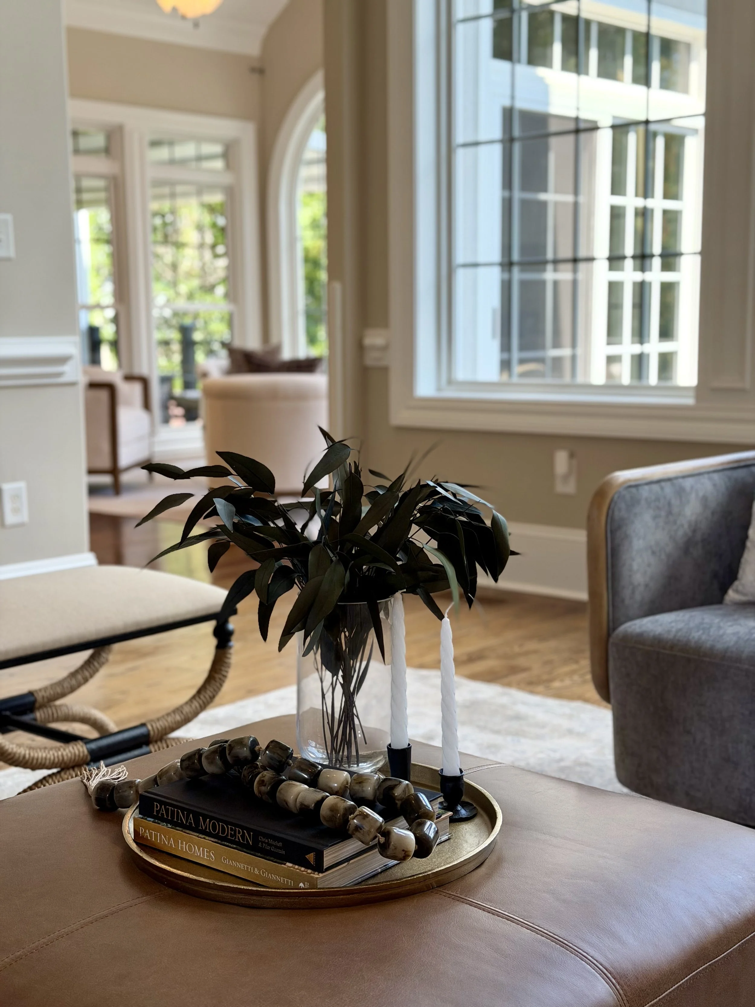 Interior view of a living room with a large window, a beige sofa, a gray armchair, and a decorative tray with books, candles, a plant, and jewelry on a table.
