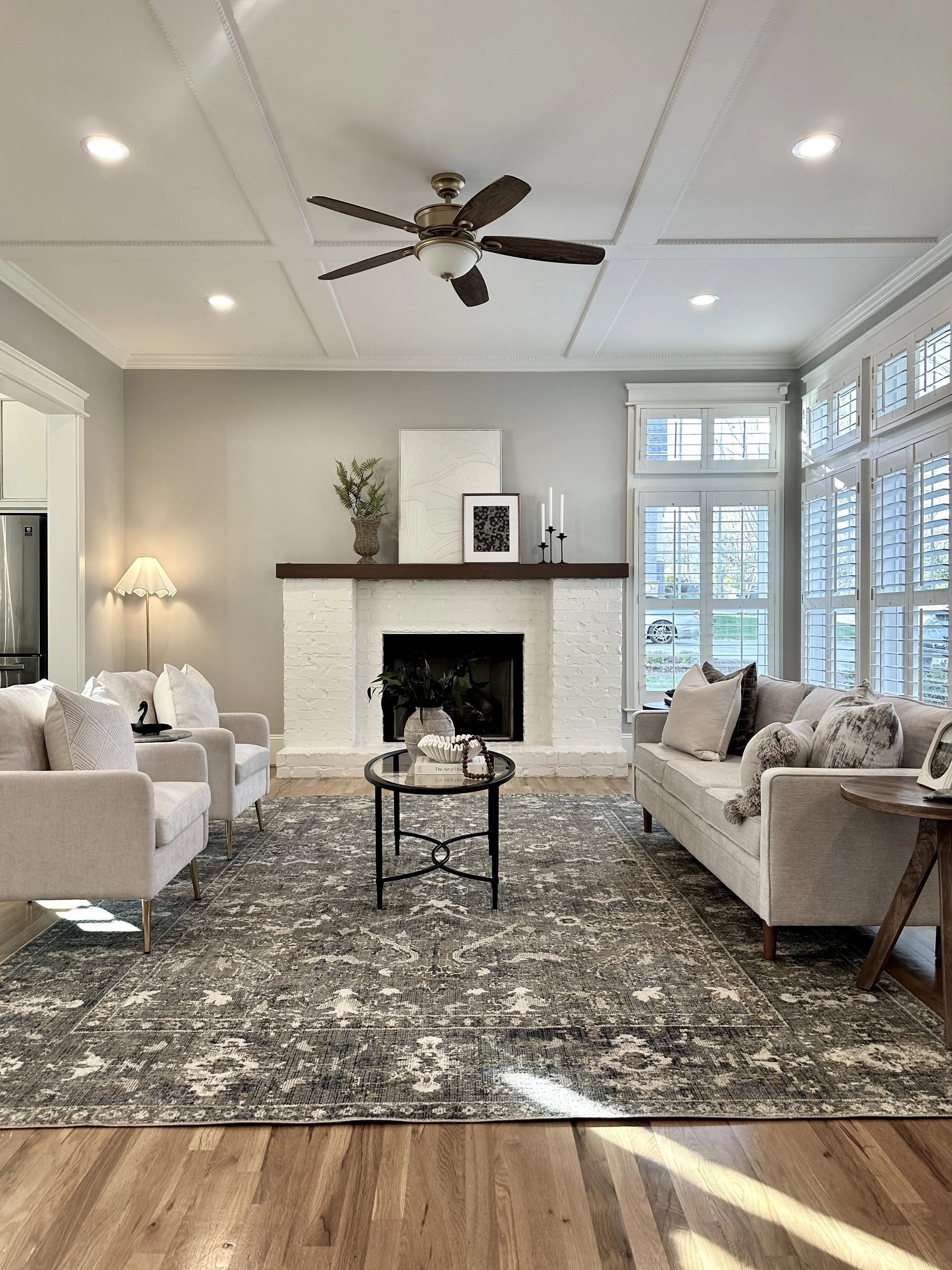 Living room with white brick fireplace, two light-colored sofas, a round black coffee table, a large patterned rug, potted plant, framed wall art, windows with white shutters, a floor lamp, and a ceiling fan.