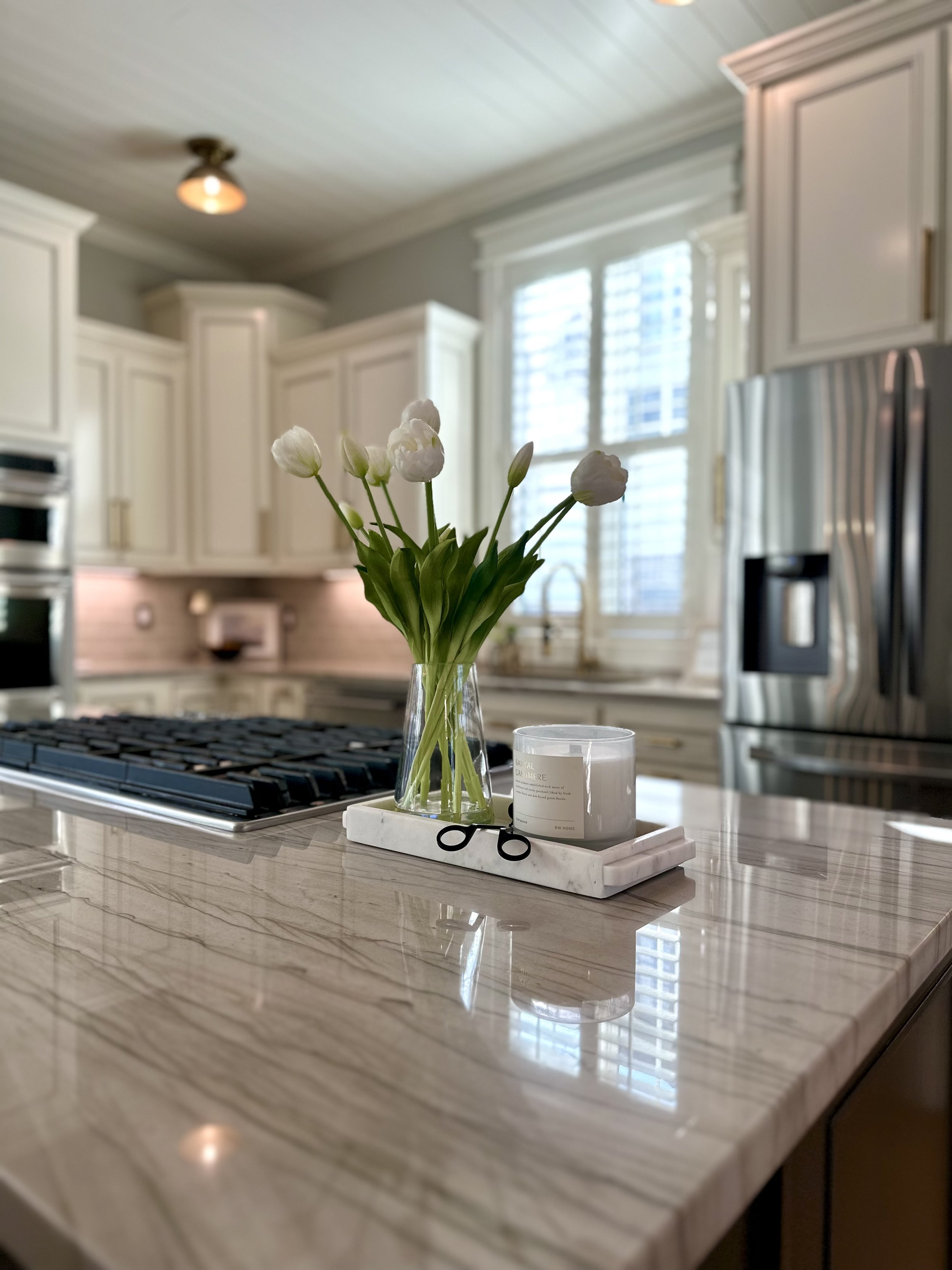 A kitchen countertop with a small vase of white flowers and a candle, with white cabinets in the background and a window with shutters.