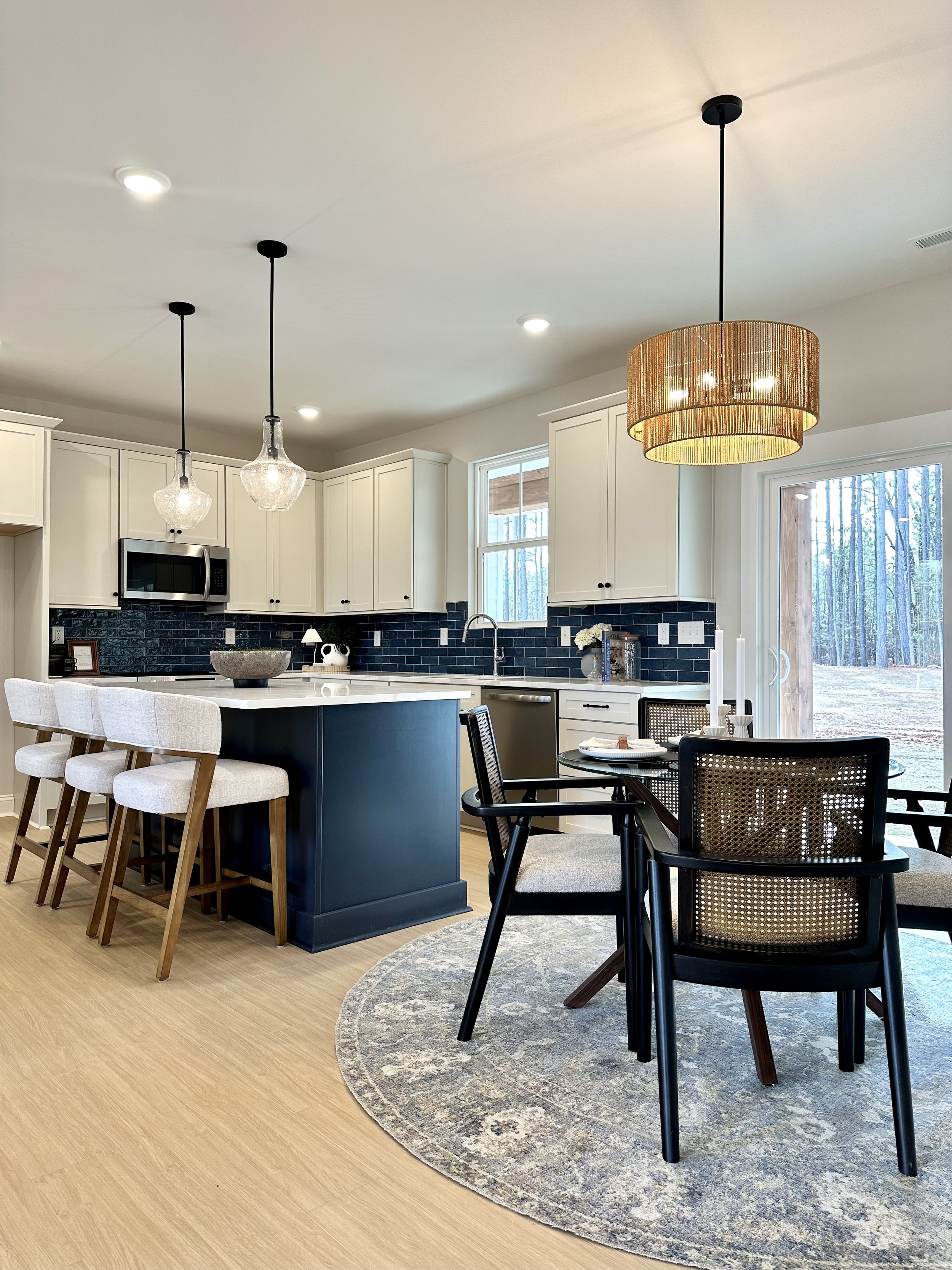Modern kitchen with white cabinets, black backsplash, island with navy blue base, and hanging pendant lights. Adjacent dining area with black chairs, a round table, and decorative candles. Large window and sliding glass door showing outdoor scenery.