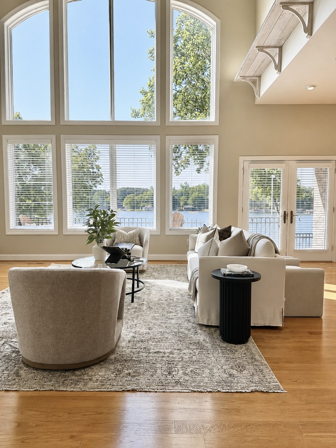 Bright living room with large windows and sliding glass door overlooking water, furnished with white sofa, armchair, coffee table, and side table, with a view of trees and water outside.