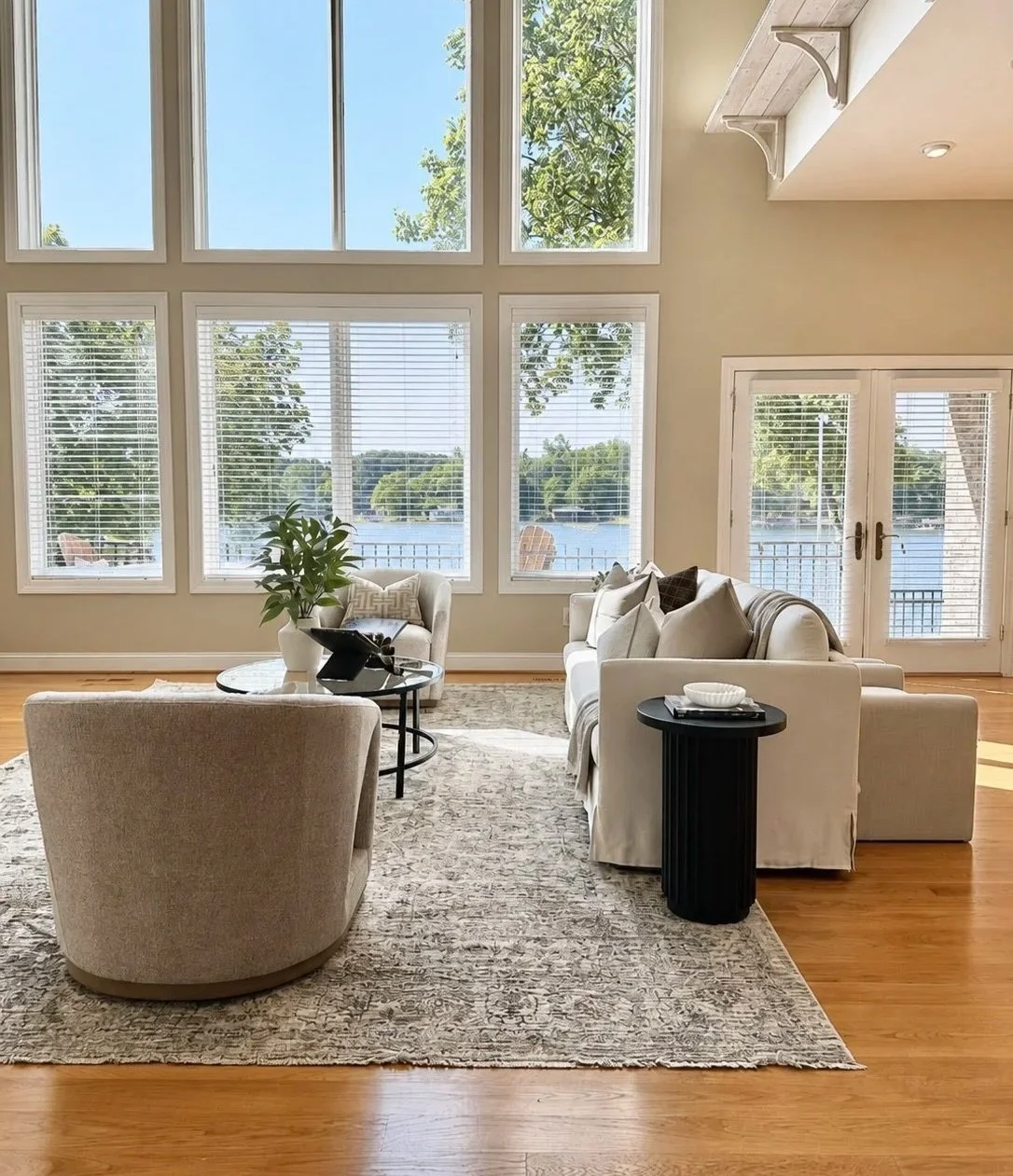 Living room with large windows overlooking a lake, featuring a white sofa, armchair, black side table, and a potted plant on a round coffee table, with hardwood flooring and a beige patterned rug.