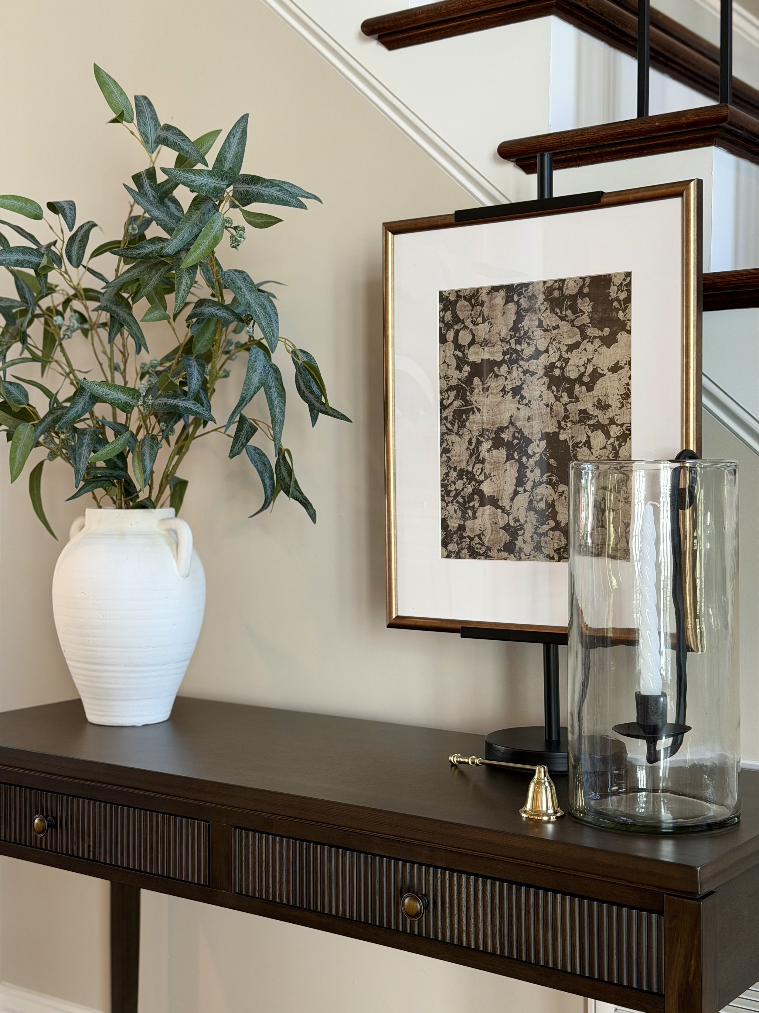 A wooden sideboard with a dark finish, featuring a large white ceramic vase with a green leafy plant, and a framed artwork with floral patterns. There is also a tall glass and a brass-colored decorative object on top of the sideboard.