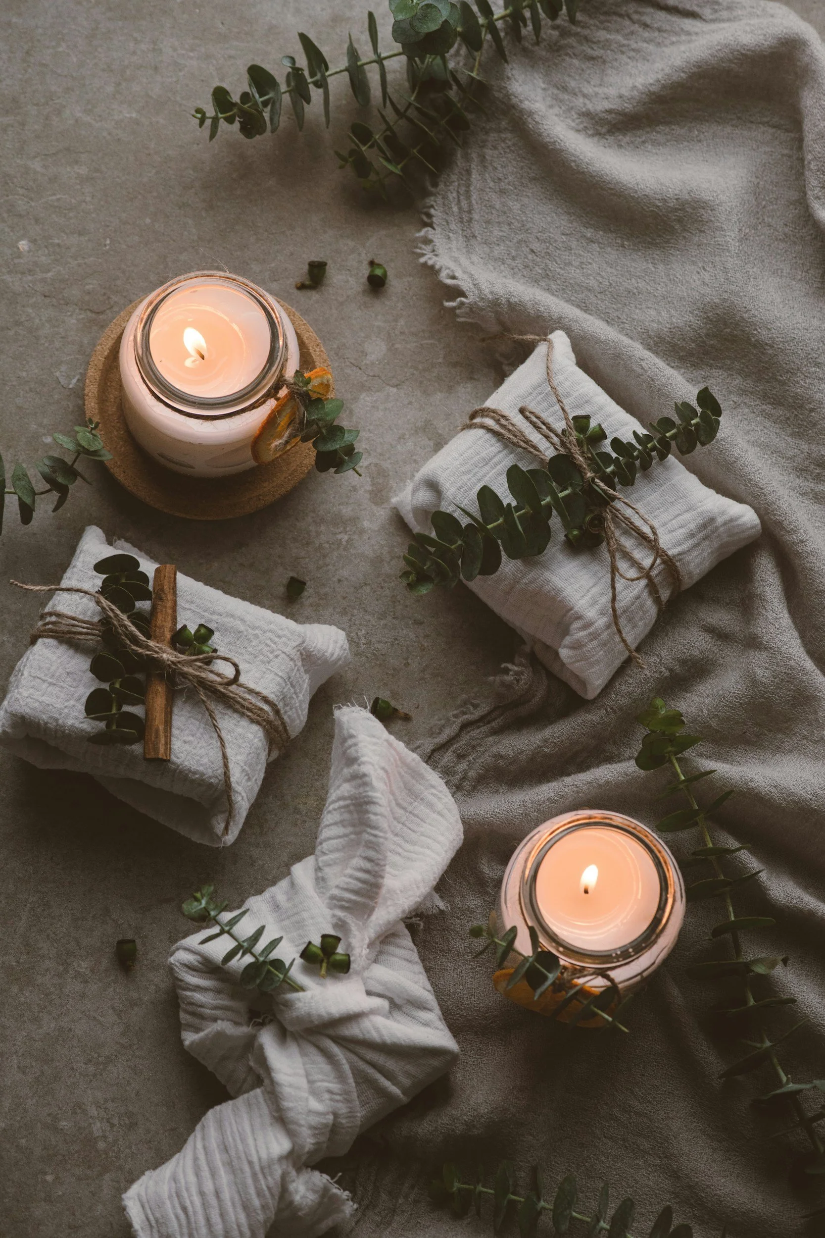 A lit candle in a glass jar on a cork coaster surrounded by greenery, linen cloth, and wrapped gifts tied with twine and decorated with small eucalyptus branches, set on a textured surface.