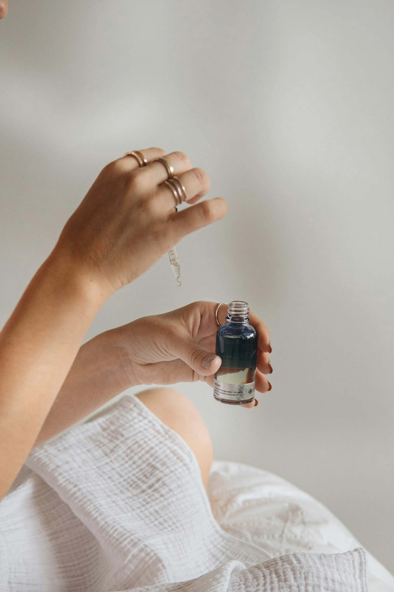 A hand with multiple rings holds a dropper above a small bottle of facial serum against a plain white wall background.