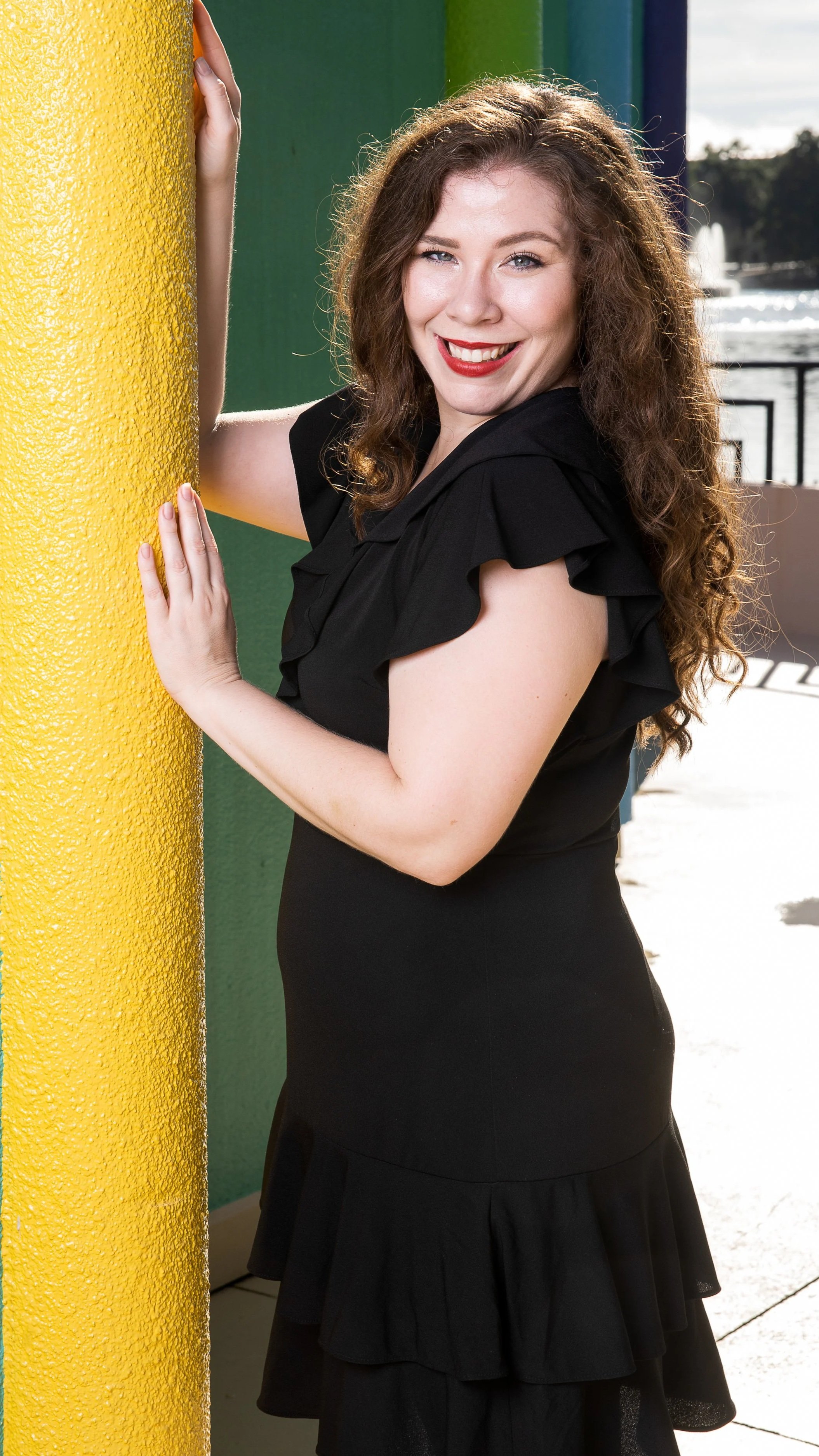 A woman with long, curly brown hair smiling and winking, wearing a black dress with ruffled sleeves, standing next to a yellow-painted pole outdoors near a body of water with fountains in the background.